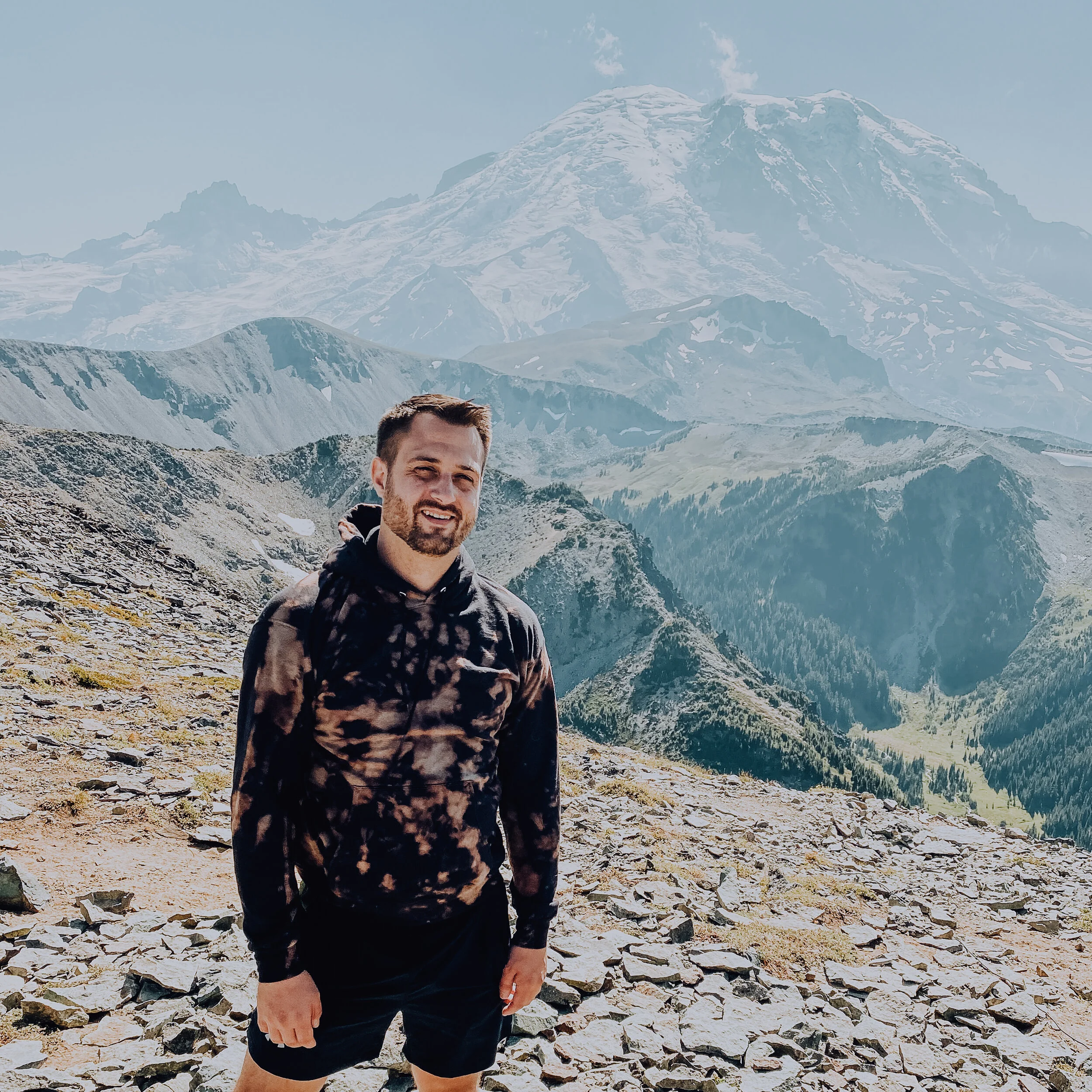 A photo of B. standing on a mountain side in a sweatshirt with snow-capped mountains behind and to the right.