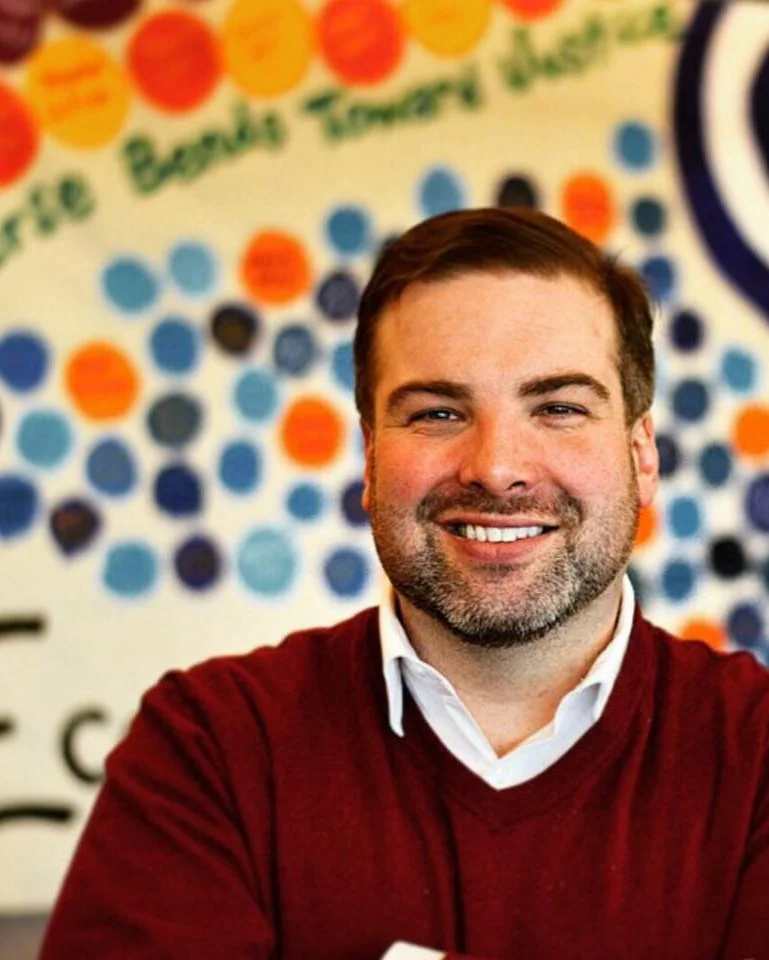 A photo of Ben in a red sweater and white button-down in front of a painted wall.