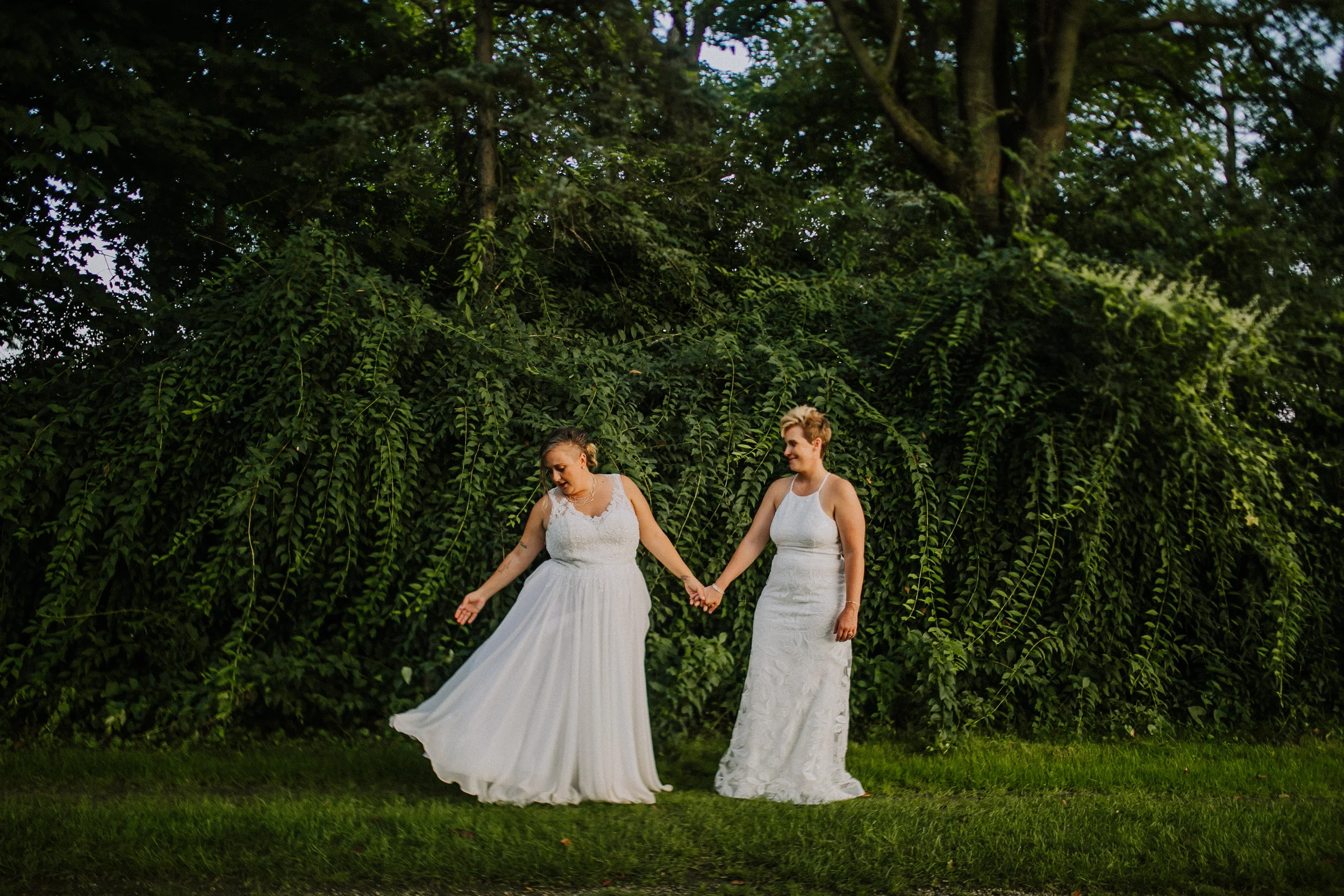 A photo of Brittany and her partner taken during their wedding. Both are in wedding dresses, holding hands, standing in front of a dense wall of green foliage.