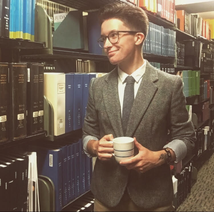 A photo of Caitlin in a tweed jacket with a tie, holding a cup of coffee, standing in a library's row of books.