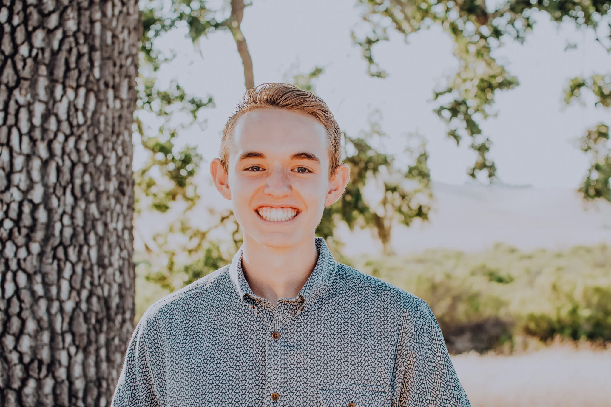 A photo of Colby in a button-down standing in front of a sunset landscape with a tree to his right.