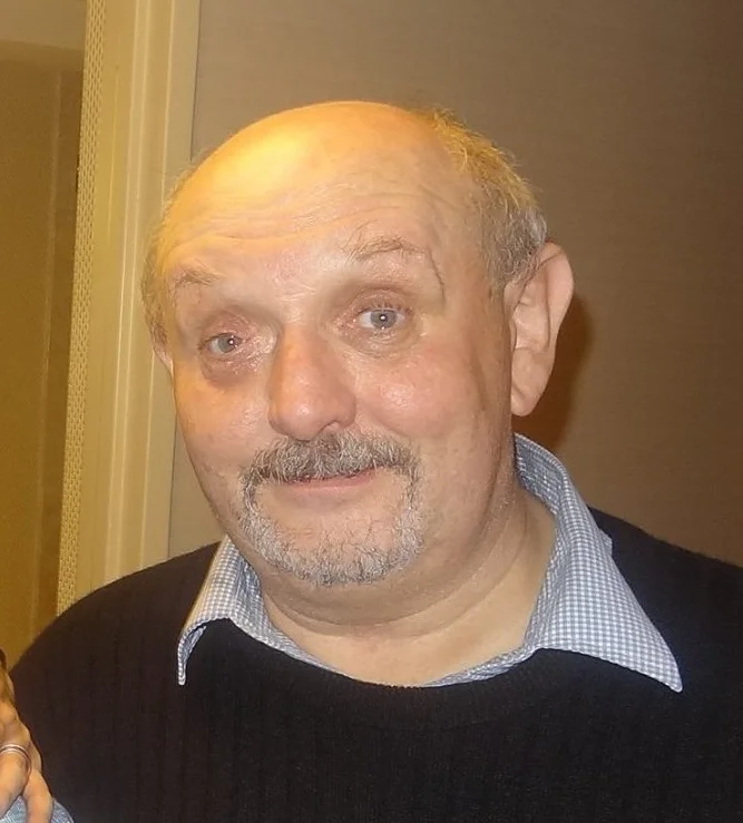 A photo of Colin in a black sweater and checkered shirt with a blank wall in the background.