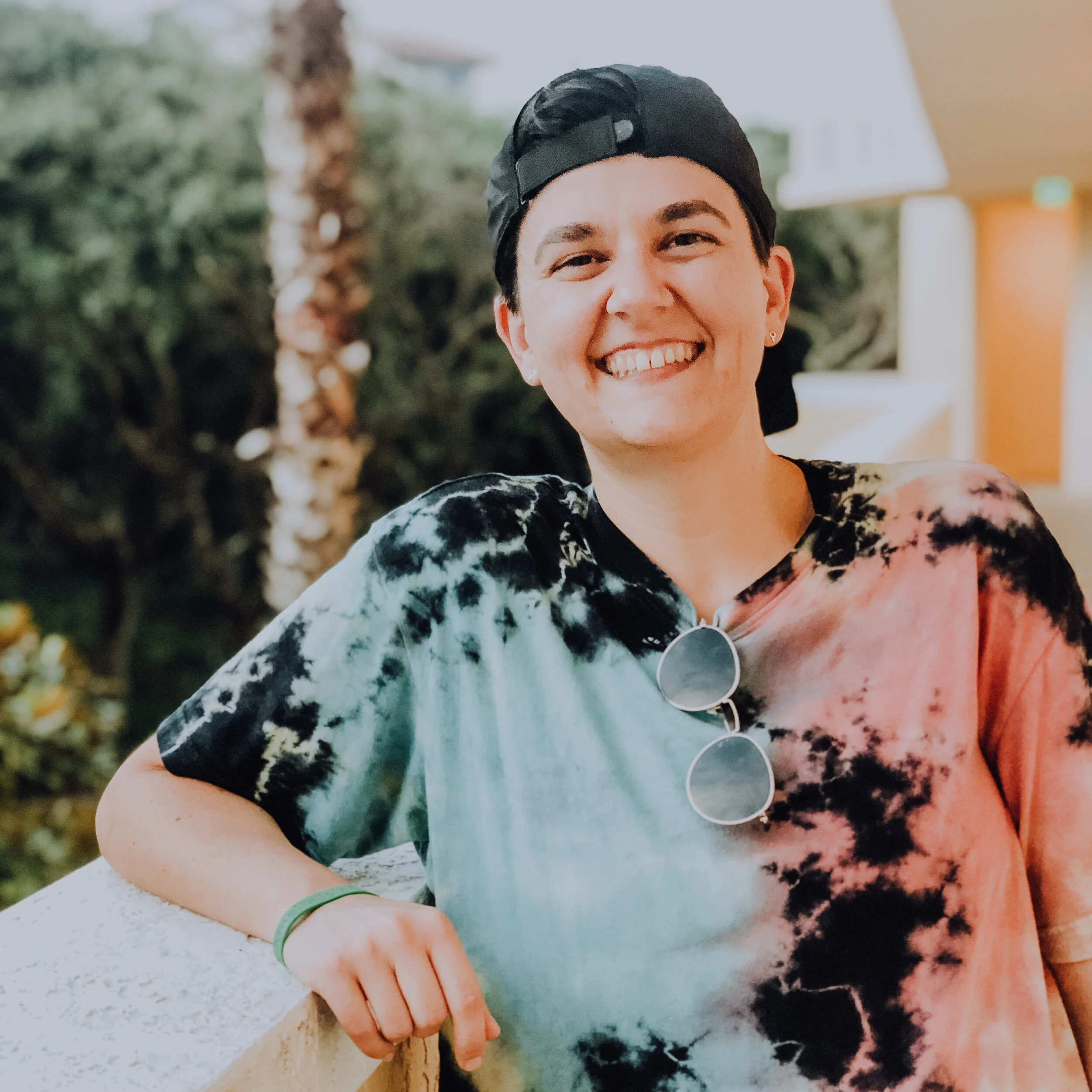 A photo of Corinne in a tie-dye shirt leaning against a railing with trees in the background.
