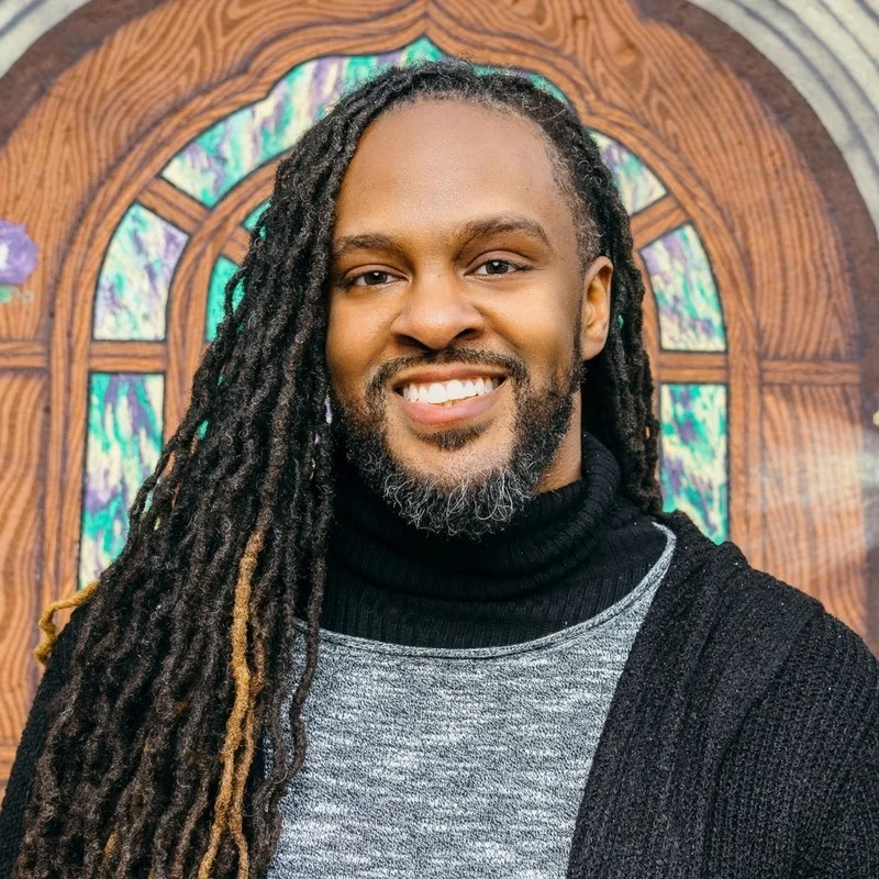 A photo of Darren in a black sweater standing in front of an illustrated stained glass window and door.