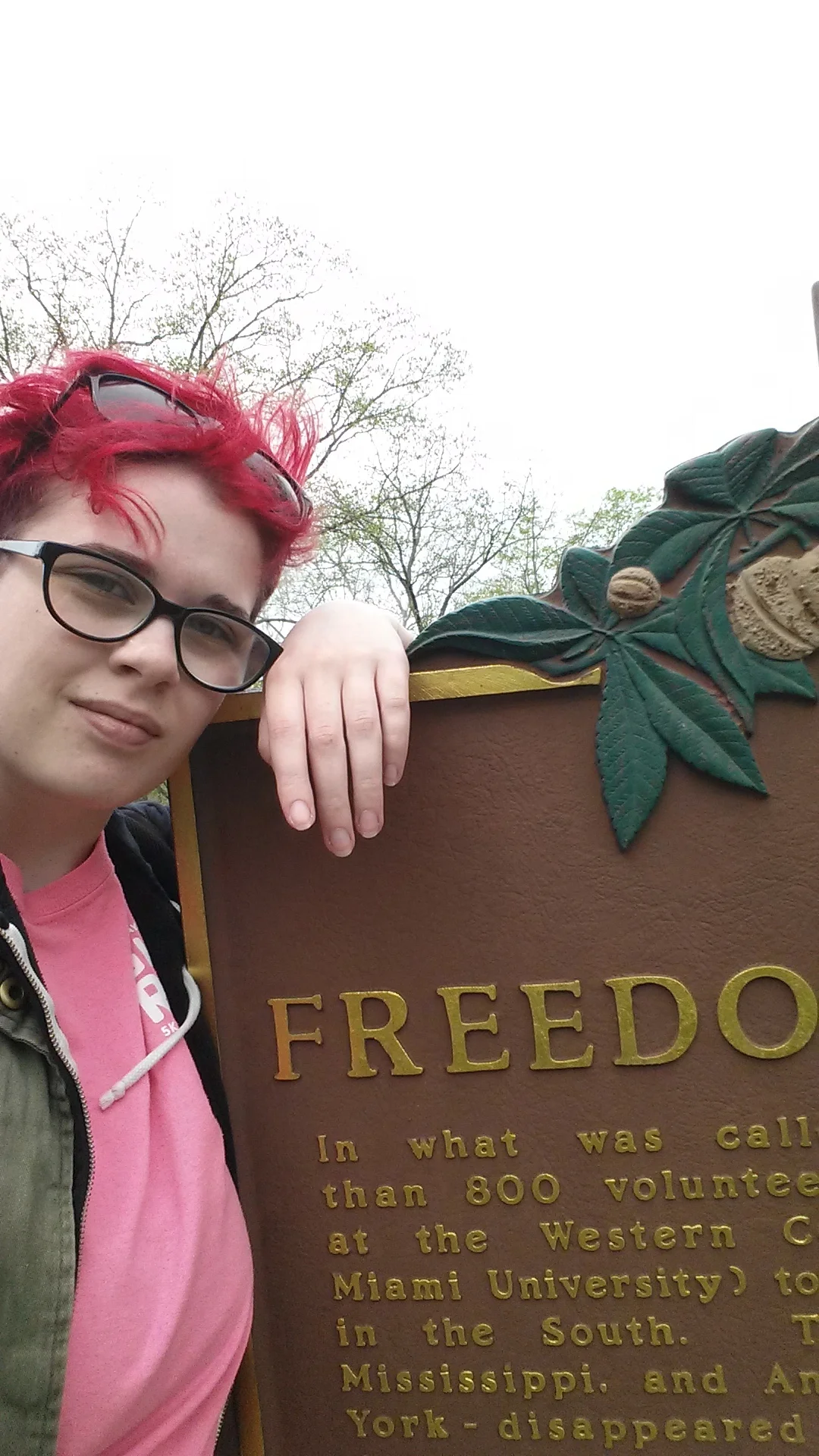 A photo of Elaine with red-pink hair leaning against a historical sign which appears to begin with the word "Freedom."