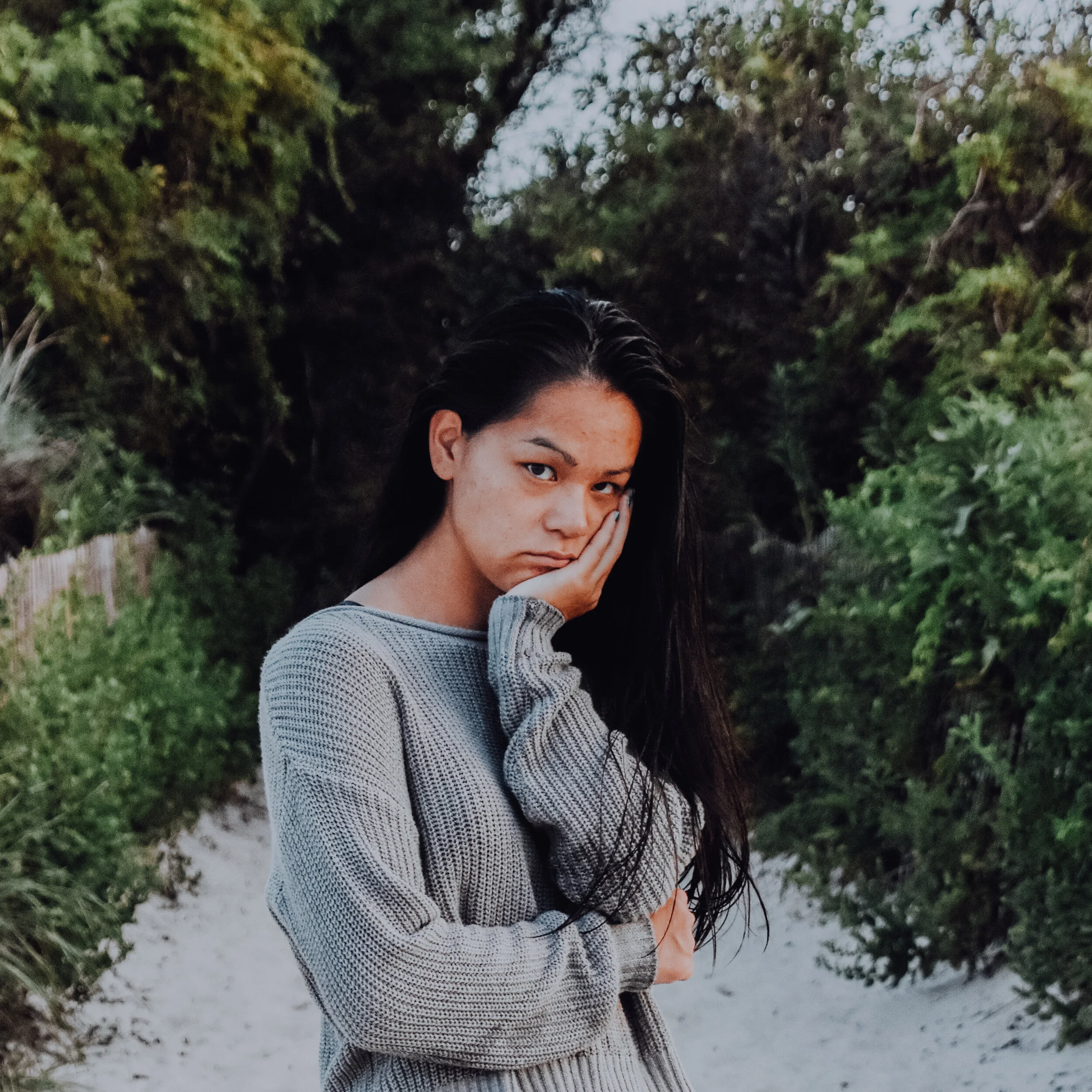A photo of Elizabeth standing on a rocky path wearing a grey sweater. There are plants to the side and behind.