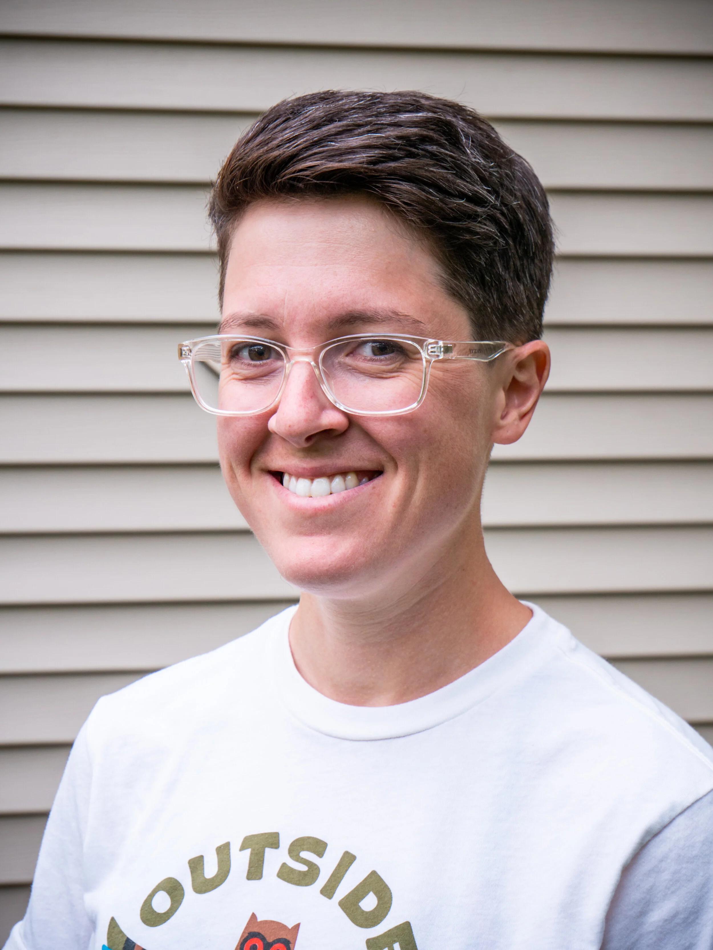 A photo of Elizabeth smiling in a white t-shirt while standing in front of beige siding.
