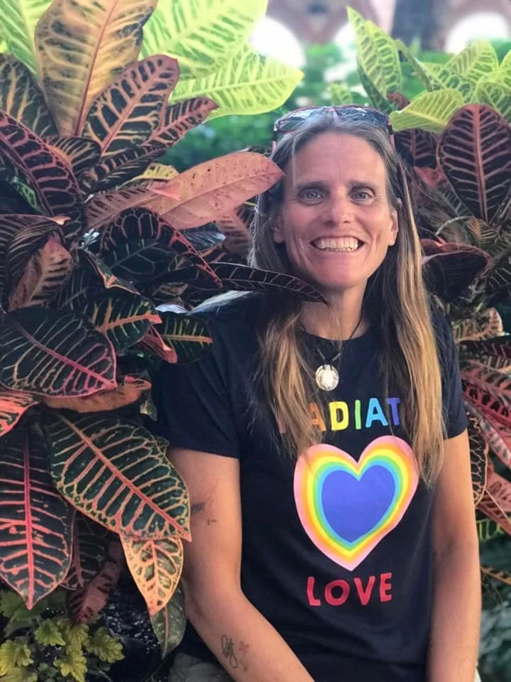A photo of Ellen standing between large plants wearing a black t-shirt and smiling.