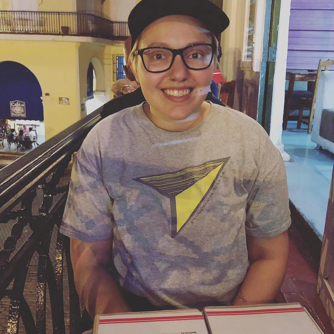 A photo of Emily sitting at a table in a cafe wearing a cap and grey t-shirt.