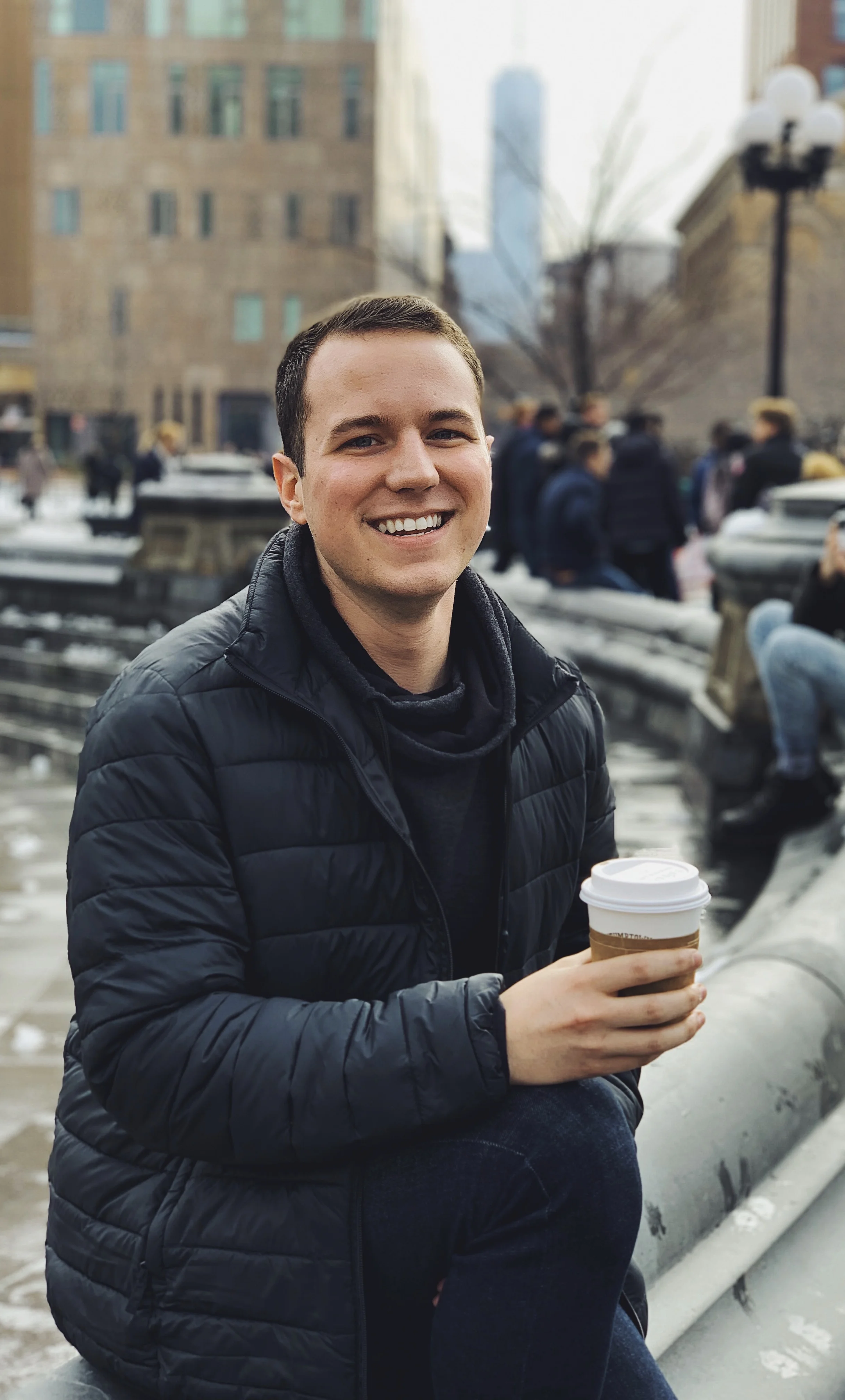 A photo of Ethan wearing a winter coat sitting against the edge of a fountain in a city. He is holding a disposable coffee cup in his right hand.