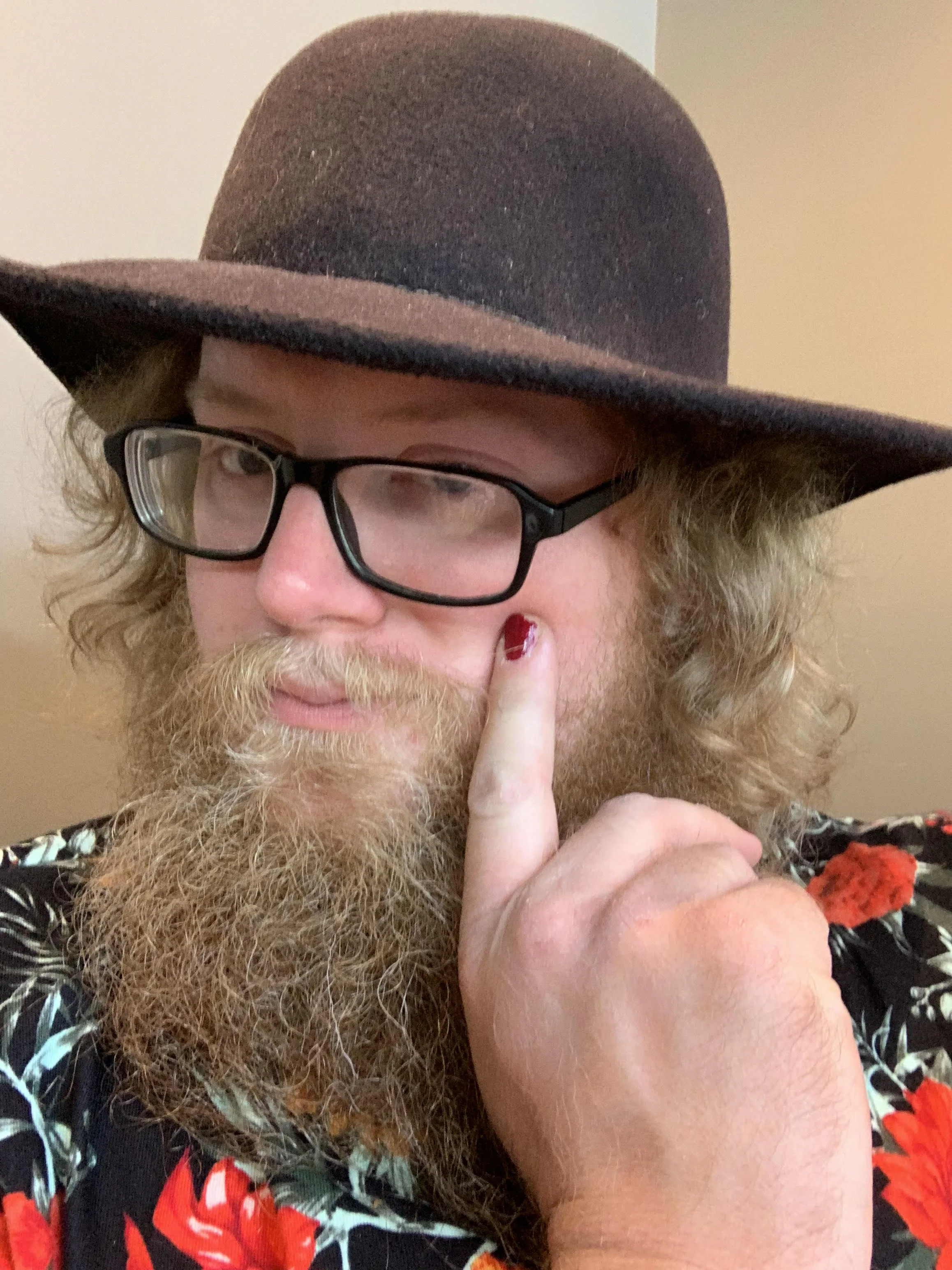 A photo of Franklyn, wearing a hat, floral shirt, and pointing a painted-fingernail towards the left eye.