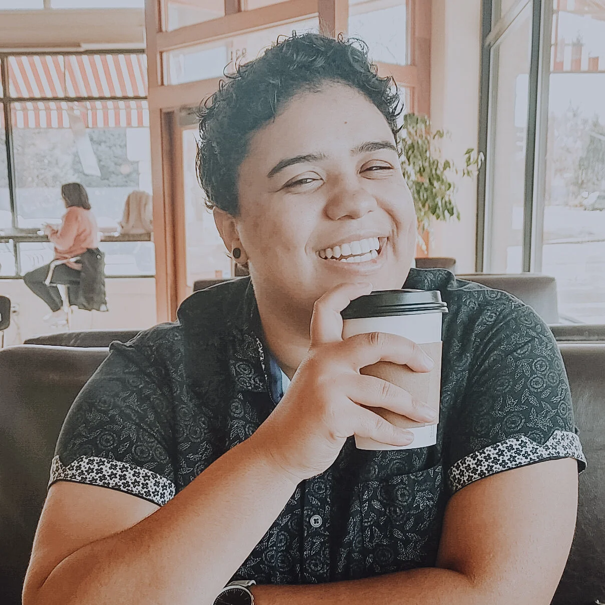 A photo of Jada smiling into the camera while sitting in a booth and drinking a cup of coffee.