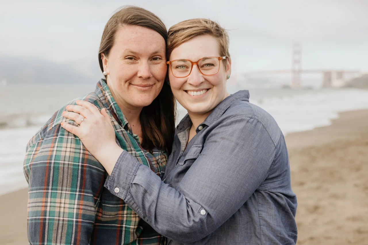 A photo of Jessica and their partner on a San Francisco beach with the fog-surrounded Golden Gate Bridge in the background.