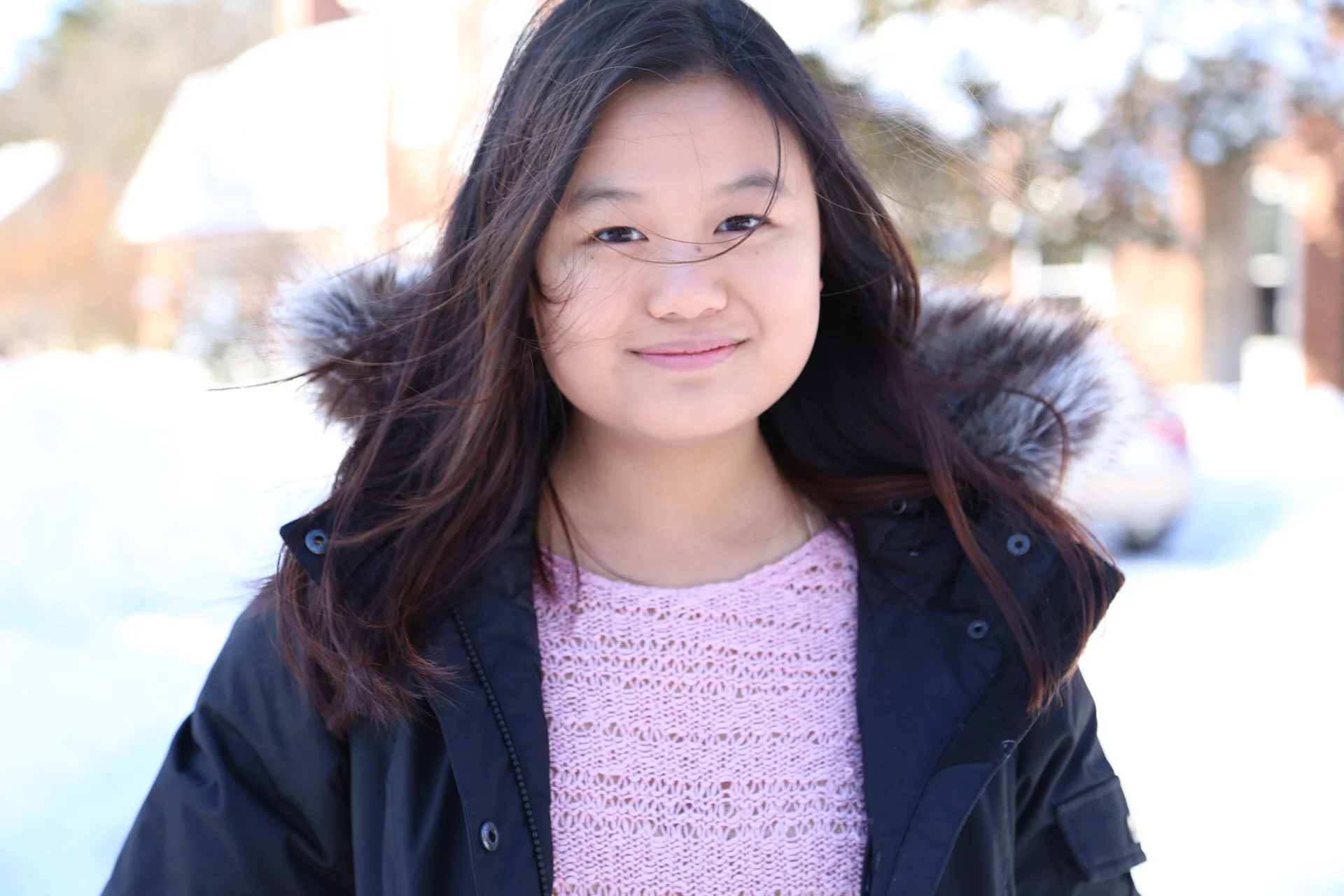 A photo of Jessica wearing a pink sweater and winter coat in front of a blurred winter landscape.