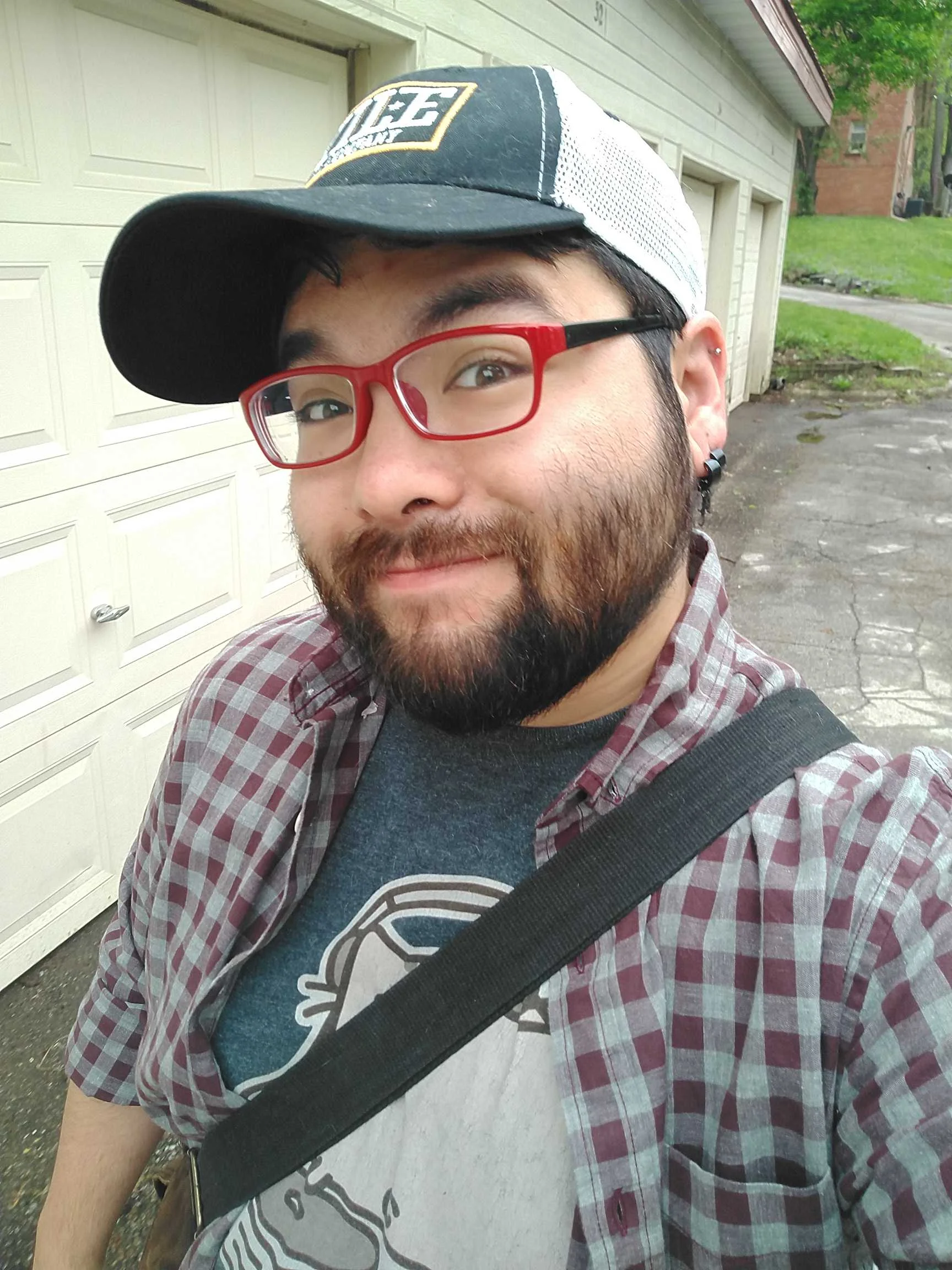 A photo of Kalevi in a checkered button-down and blue t-shirt, wearing a cap, with red-framed glasses. Kalevi is standing in front of a series of garage doors.