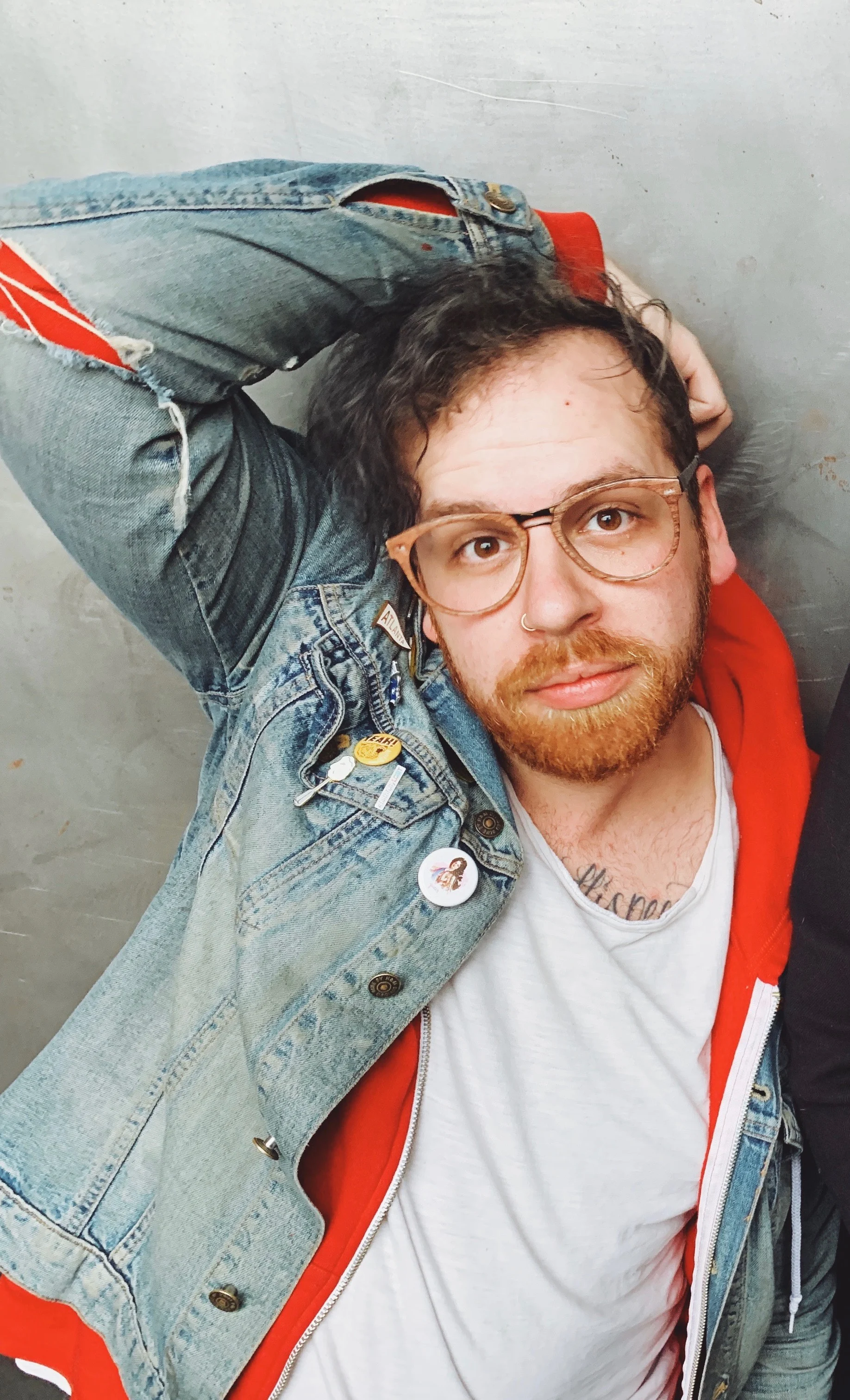 A photo of Kevin in a denim jacket, red sweatshirt, and white t-shirt leaning against a concrete call with an arm over their head.