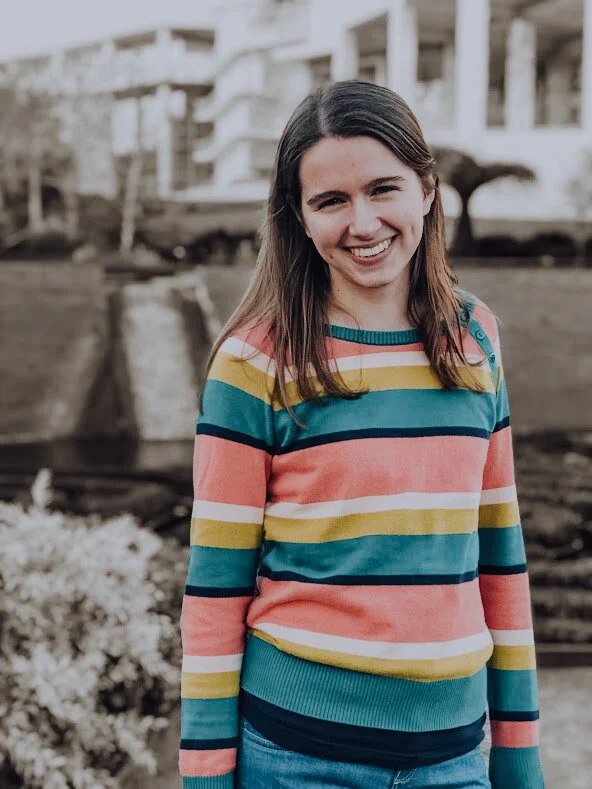 A photo of Lauren wearing a pastel-banded shirt in front of a building where the background is in neutral, sepia tones.