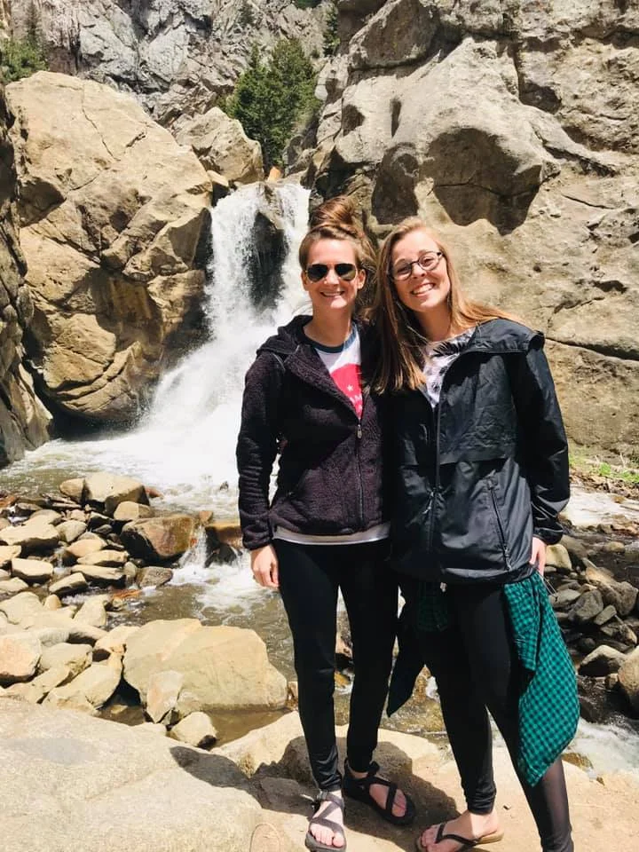A photo of Lauren and her partner Hannah standing in front of a rocky cliff face with a waterfall in the background.
