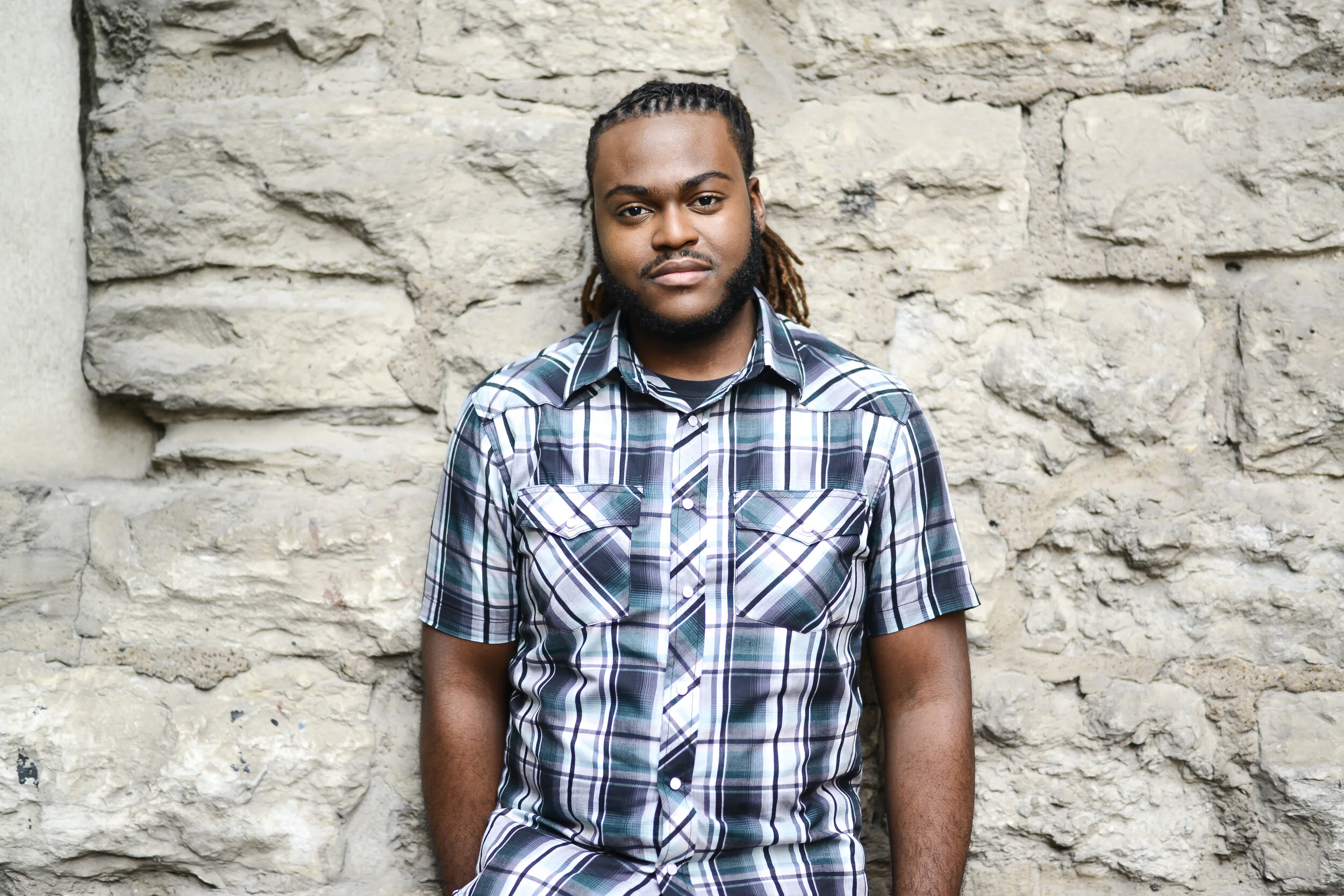 A photo of Luther standing in front of a rock wall wearing a plaid button-down shirt.