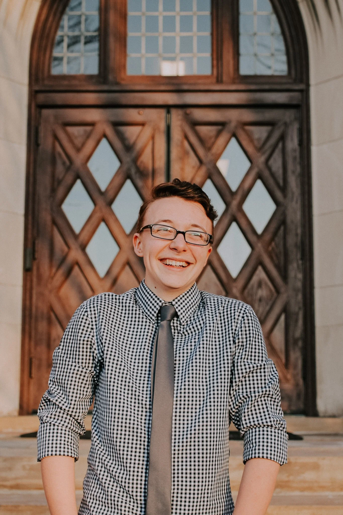 A photo of Mack in a checkered shirt and grey tie, smiling in front of a large wood and glass doorway.
