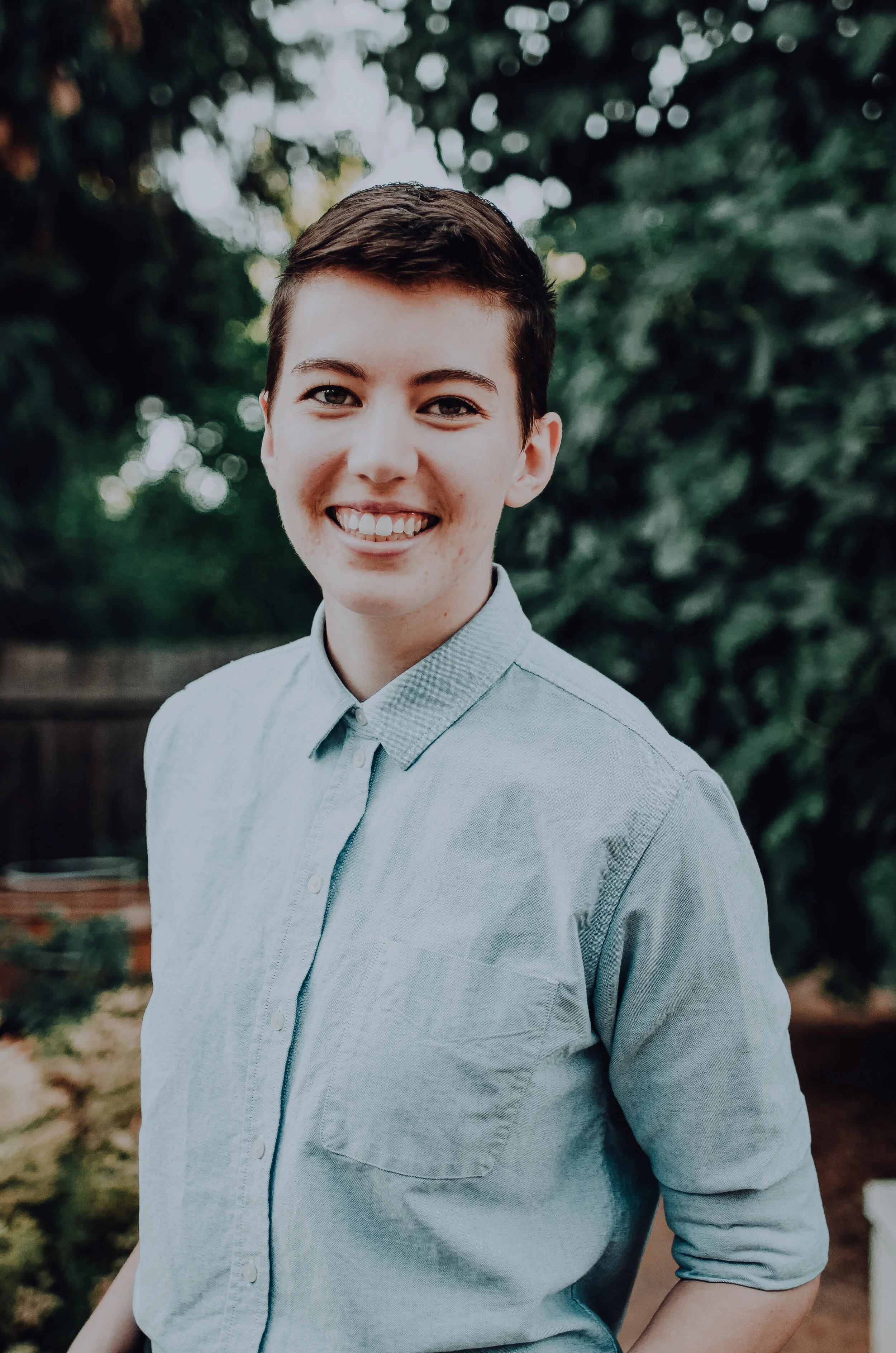 A photo of Mary in a blue button-down shirt standing in front of green foliage.