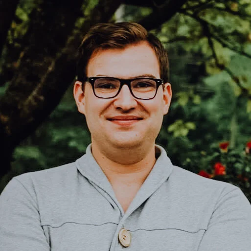 A photo of Matthew smiling in a grey sweater in front of green treen foliage.
