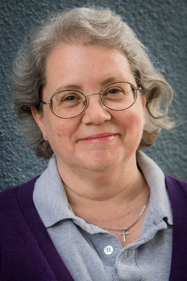 A photo of Melinda in a purple cardigan, grey polo, against a pale blue wall.