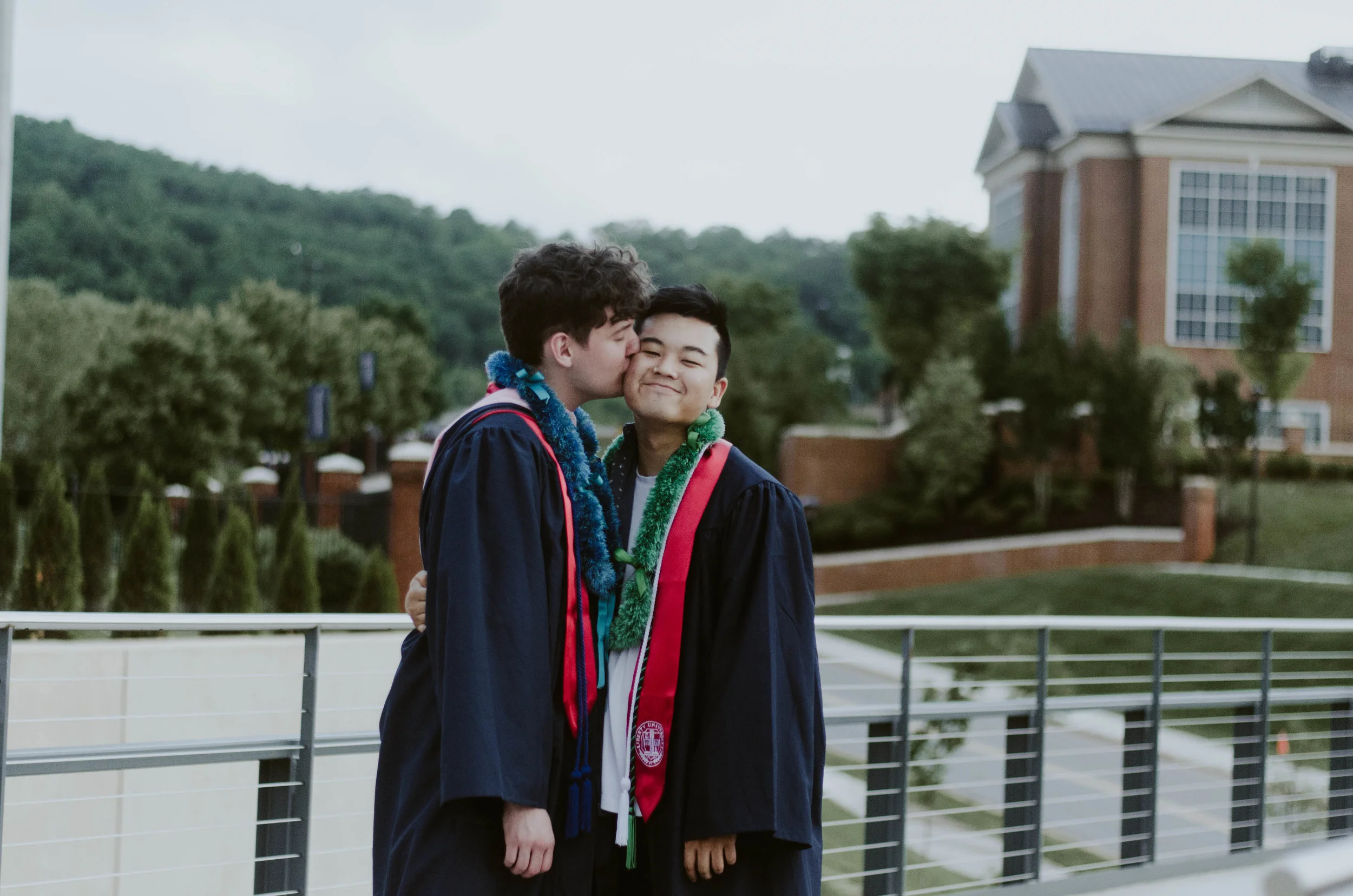A photo of Nathanial kissing his partner Elliot on a sidewalk on Liberty's campus. Both are dressed in graduation gowns.