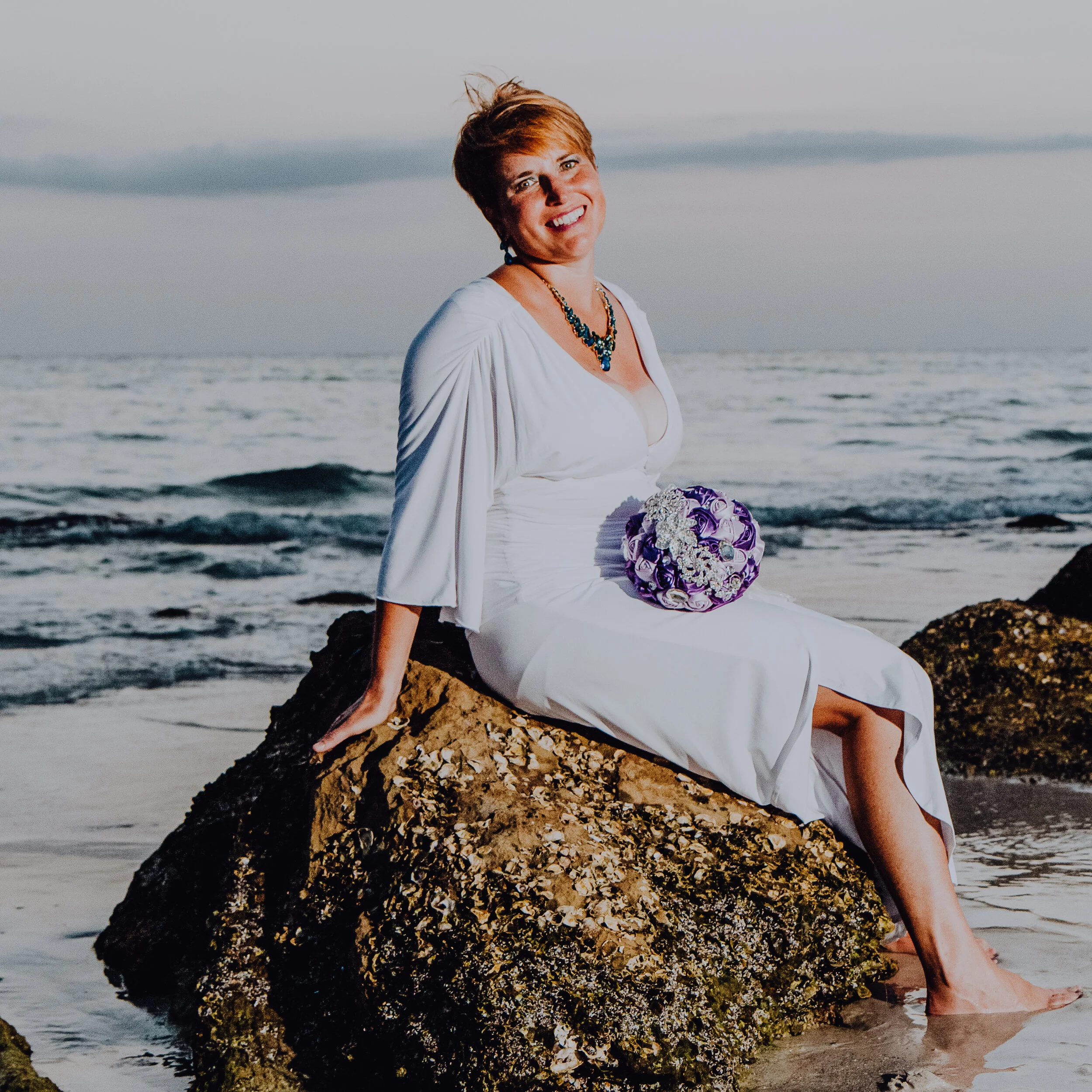 A photo of RJ sitting on a rock on the beach with a bocquet on her lap and the ocean behind her.