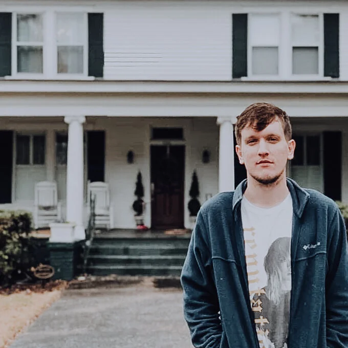 A photo of Rob in a sweatshirt standing in front of a white house porch.