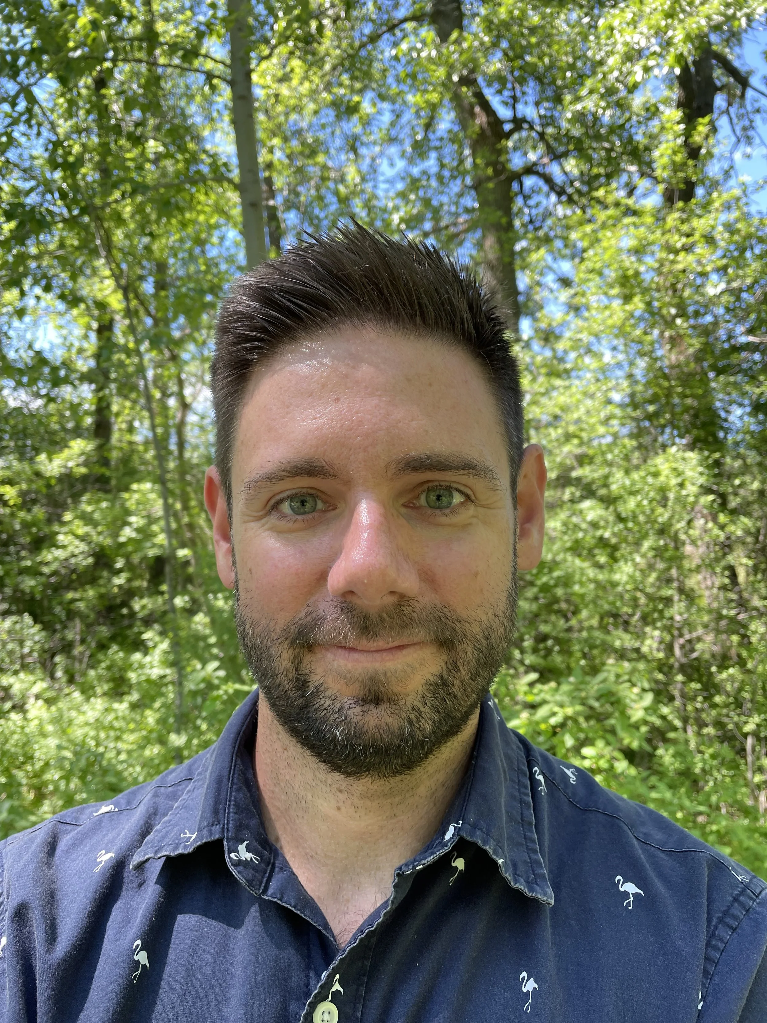 A selfie of Shane wearing a blue collared shirt in front of a treeline.