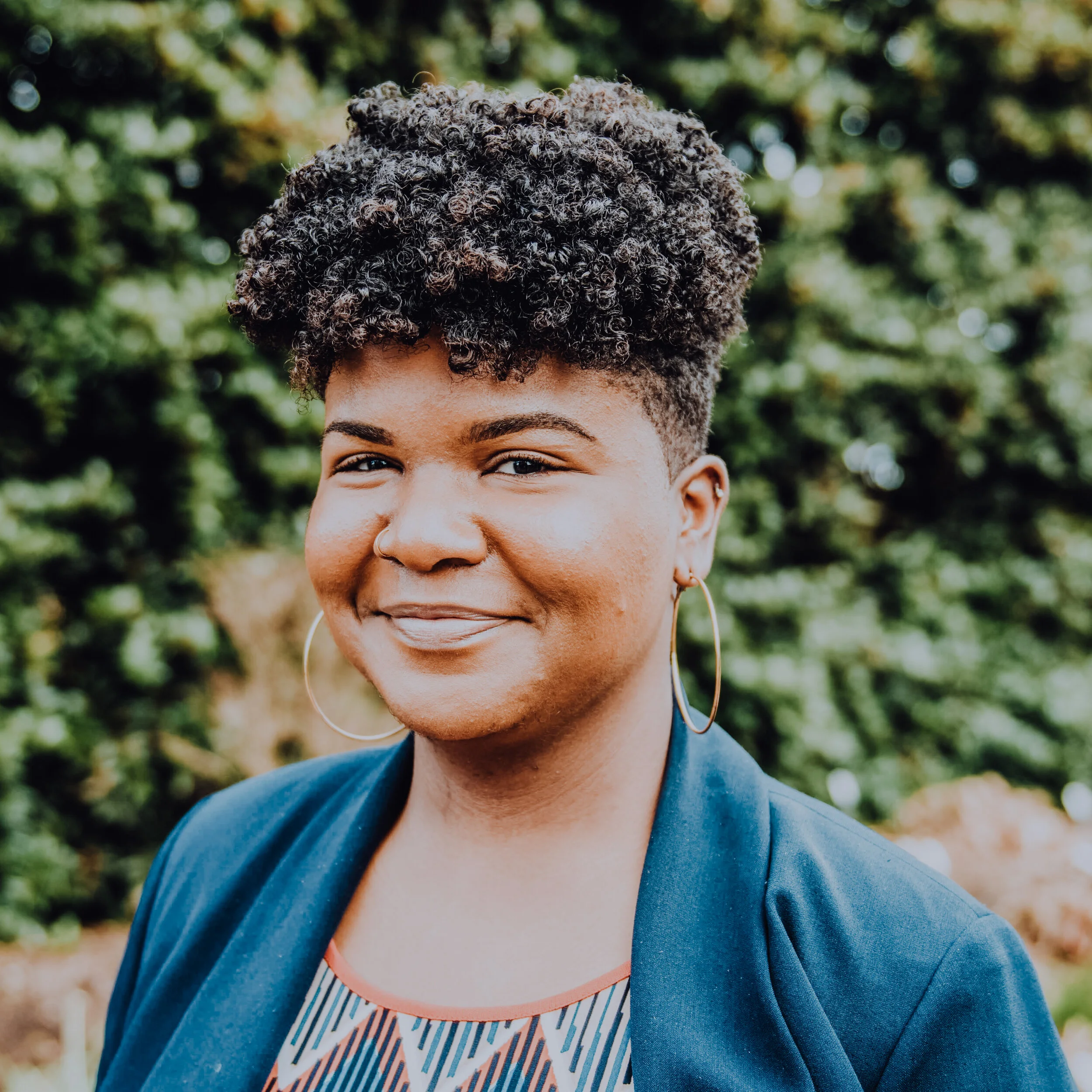 A photo of sharre wearing a blue blazer, smiling into the camera while standing in front of foliage.