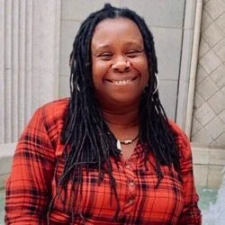 A photo of Unoma in a red plaid button-down standing in front of a stone building.