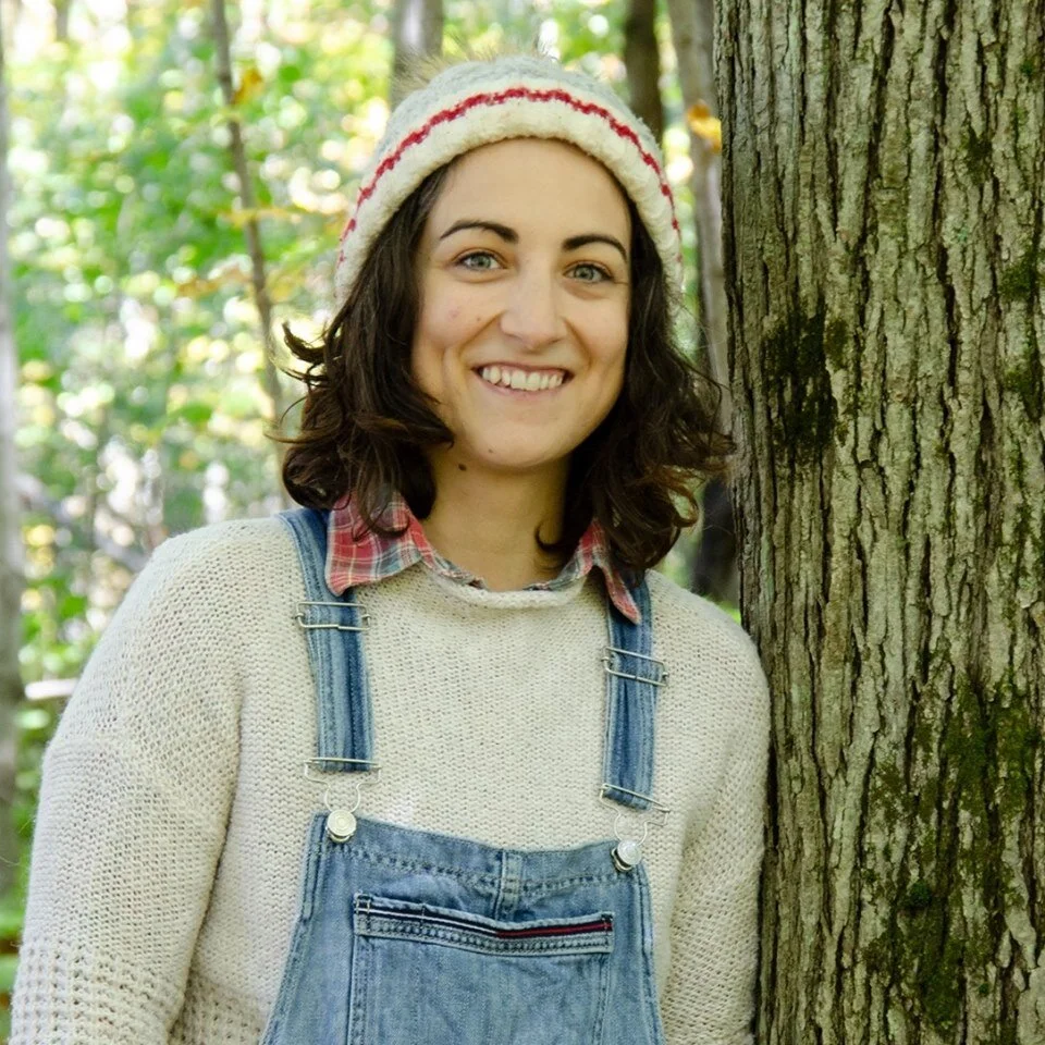 A photo of Vicky leaning against a tree in the woods wearing a sweater and denim overalls.