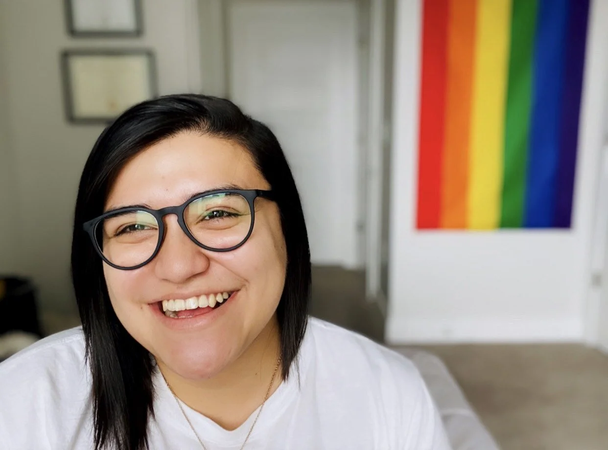 Photo of a contributor smiling with a pride flag behind them.