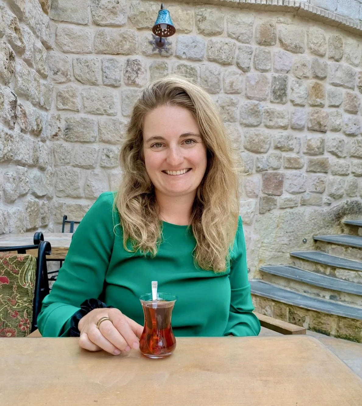 A woman with wavy blonde hair smiling and sitting at a table in an outdoor setting, holding a glass of Turkish tea, with a stone wall behind her and stairs to the side.