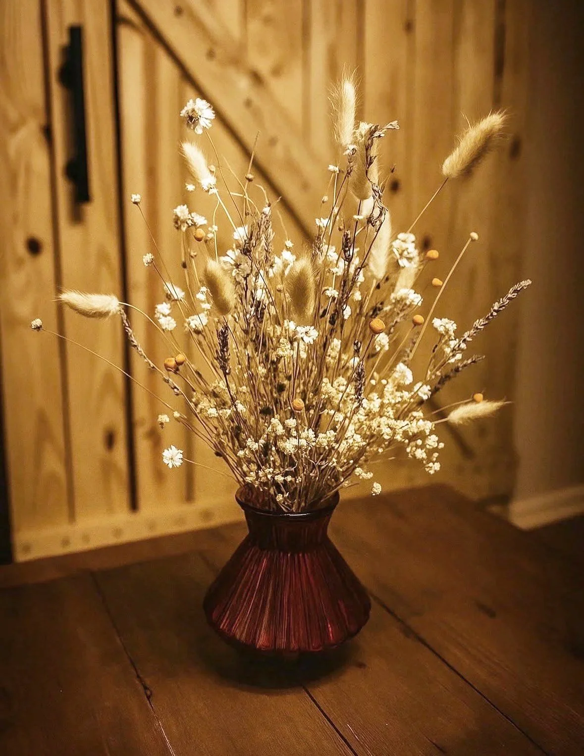 A brown glass vase filled with dried flowers and grasses, placed on a wooden table in front of a wooden wall.