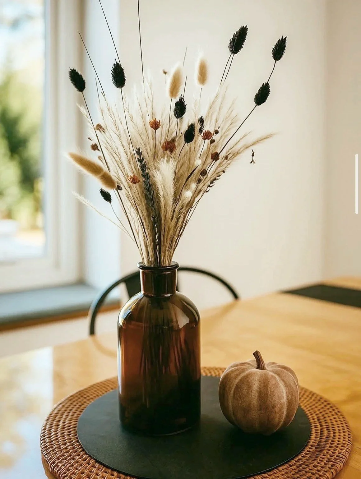 Decorative dried flower bouquet in a brown glass vase on a round woven placemat with a small artificial pumpkin on a black mat, on a wooden table near a window.