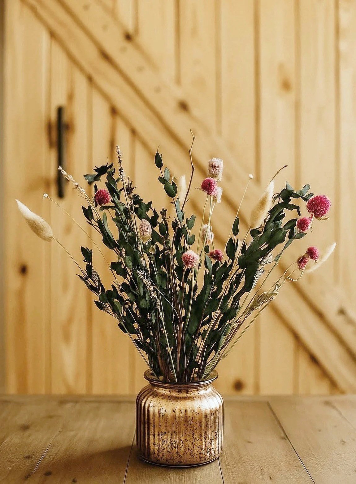 A bouquet of dried flowers with pink and white accents in a rustic ceramic vase on a wooden table against a wooden-paneled wall.