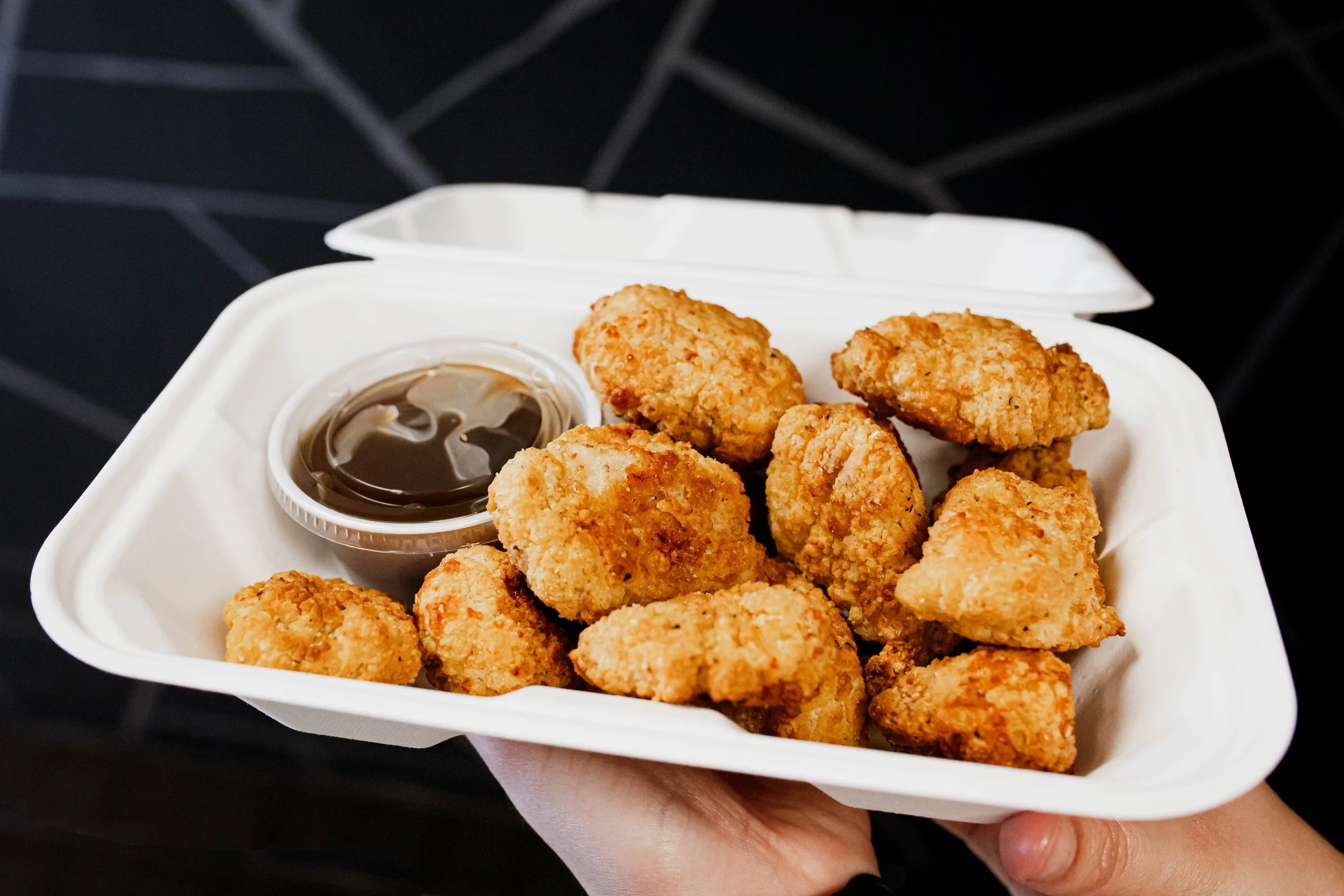 Fried chicken nuggets with a side of dark dipping sauce in a white takeout box.