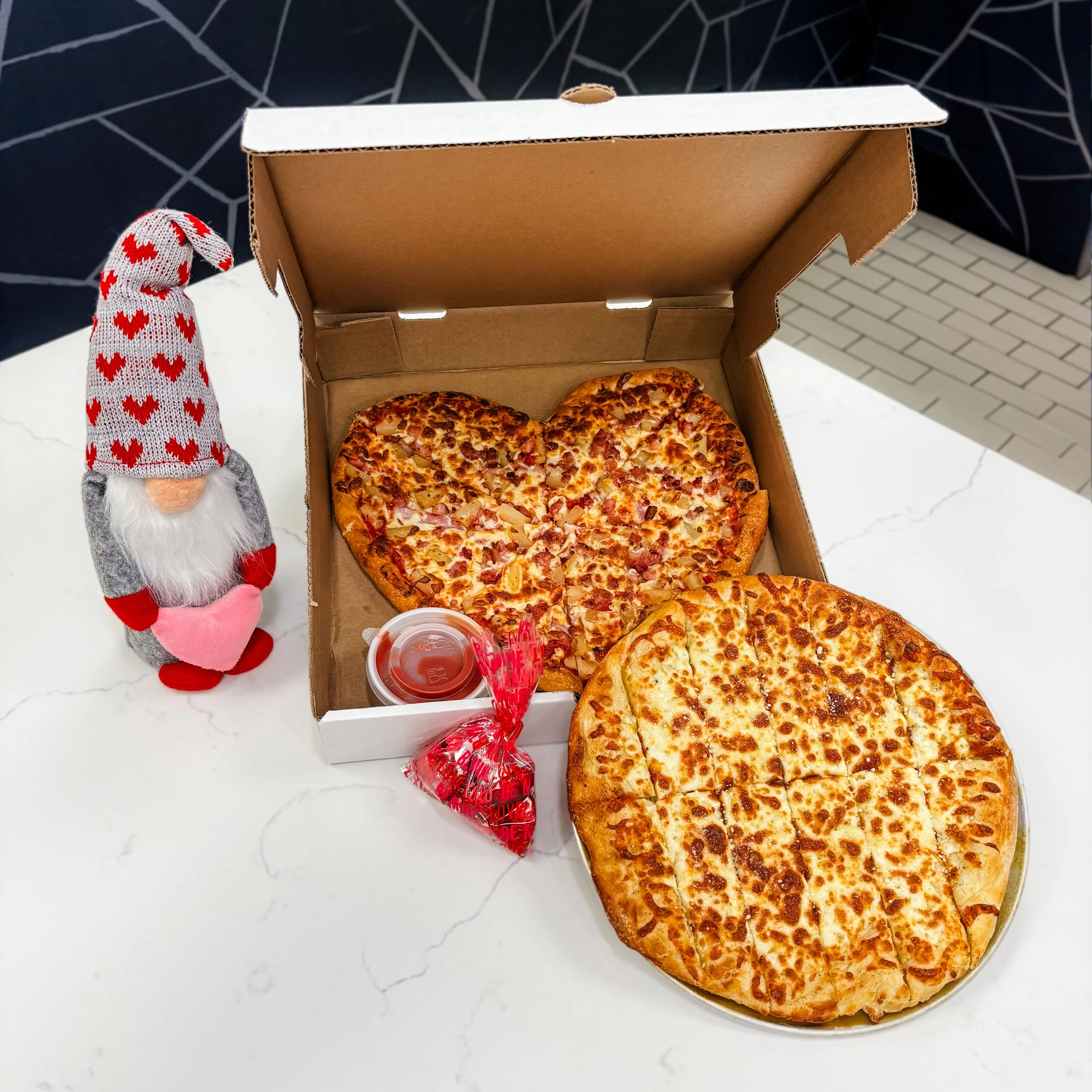 A pizza in a box with one heart-shaped half pepperoni and pineapple pizza and a large round cheese pizza on a white marble table. Next to the pizza is a small cup of sauce and a red heart-shaped bag of dipping sauce. There is also a plush gnome toy with a grey beard, red heart-patterned hat, and red shoes standing to the left of the pizza box.