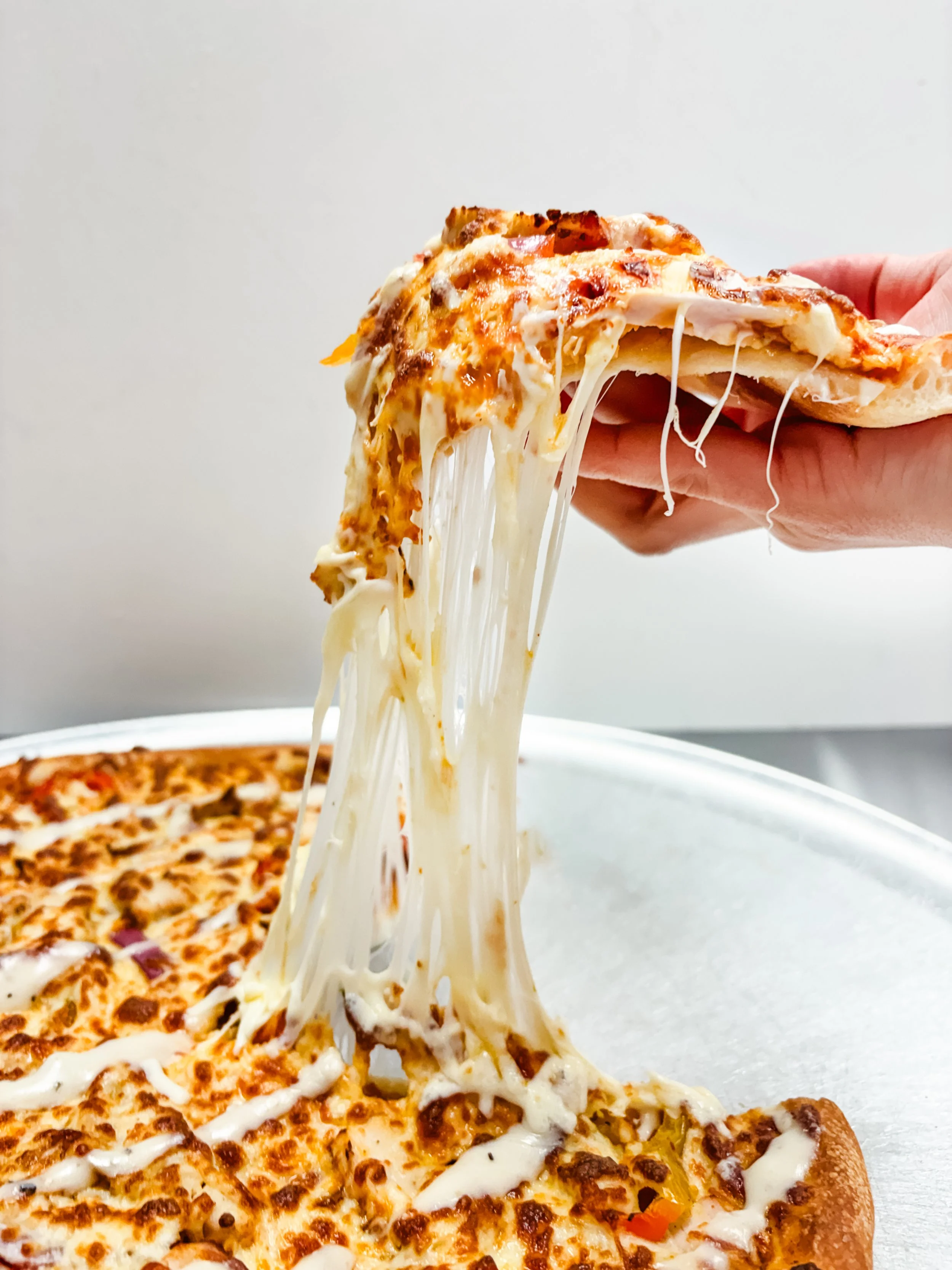 Close-up of a hand lifting a slice of cheese pizza with melted cheese stretching between the slice and the remaining pizza. The pizza has a golden-brown crust and is topped with cheese and possibly some vegetables or meat.