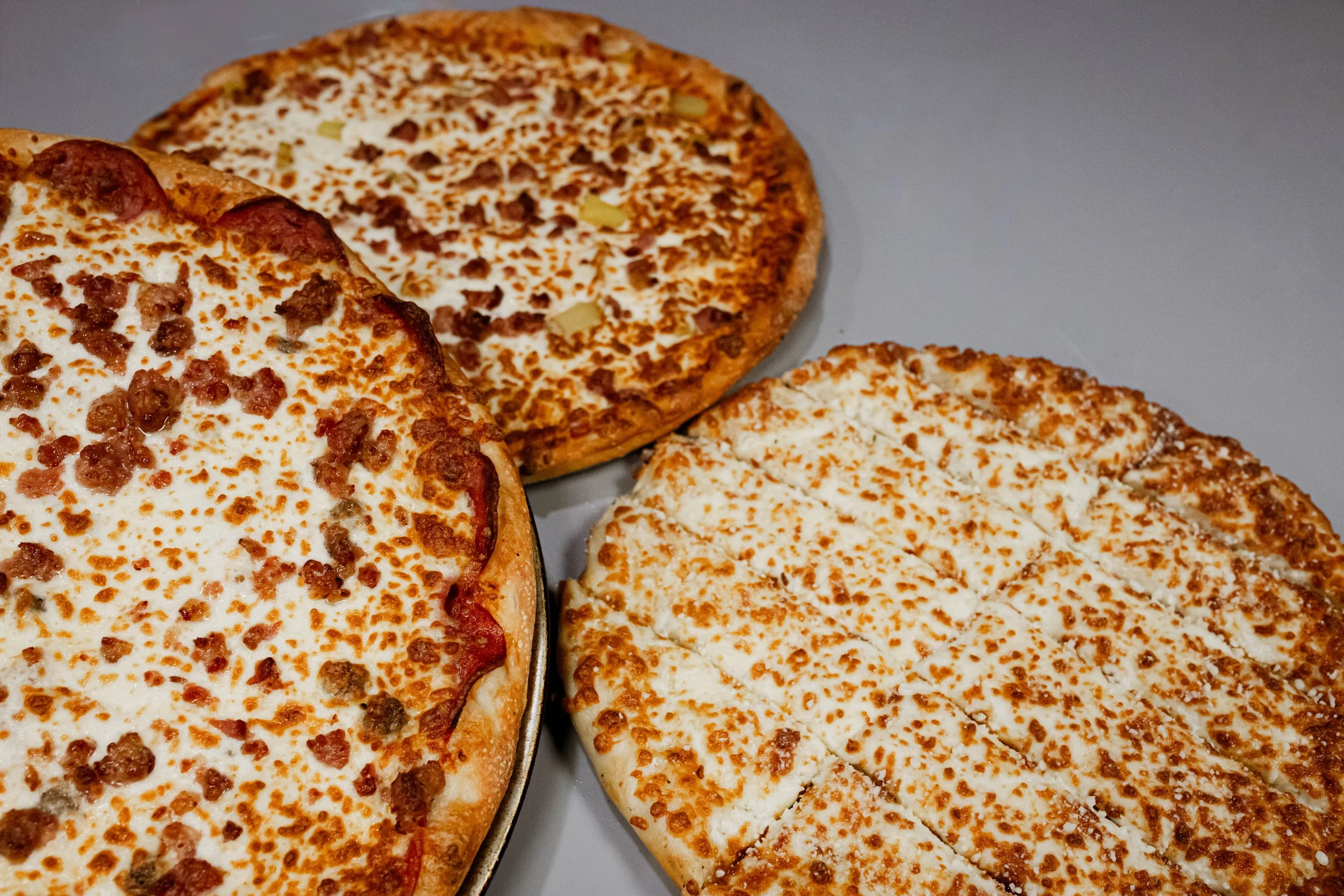 Three pizzas on a table: one cheese and sausage, one cheese and possibly pineapple, and one plain cheese.
