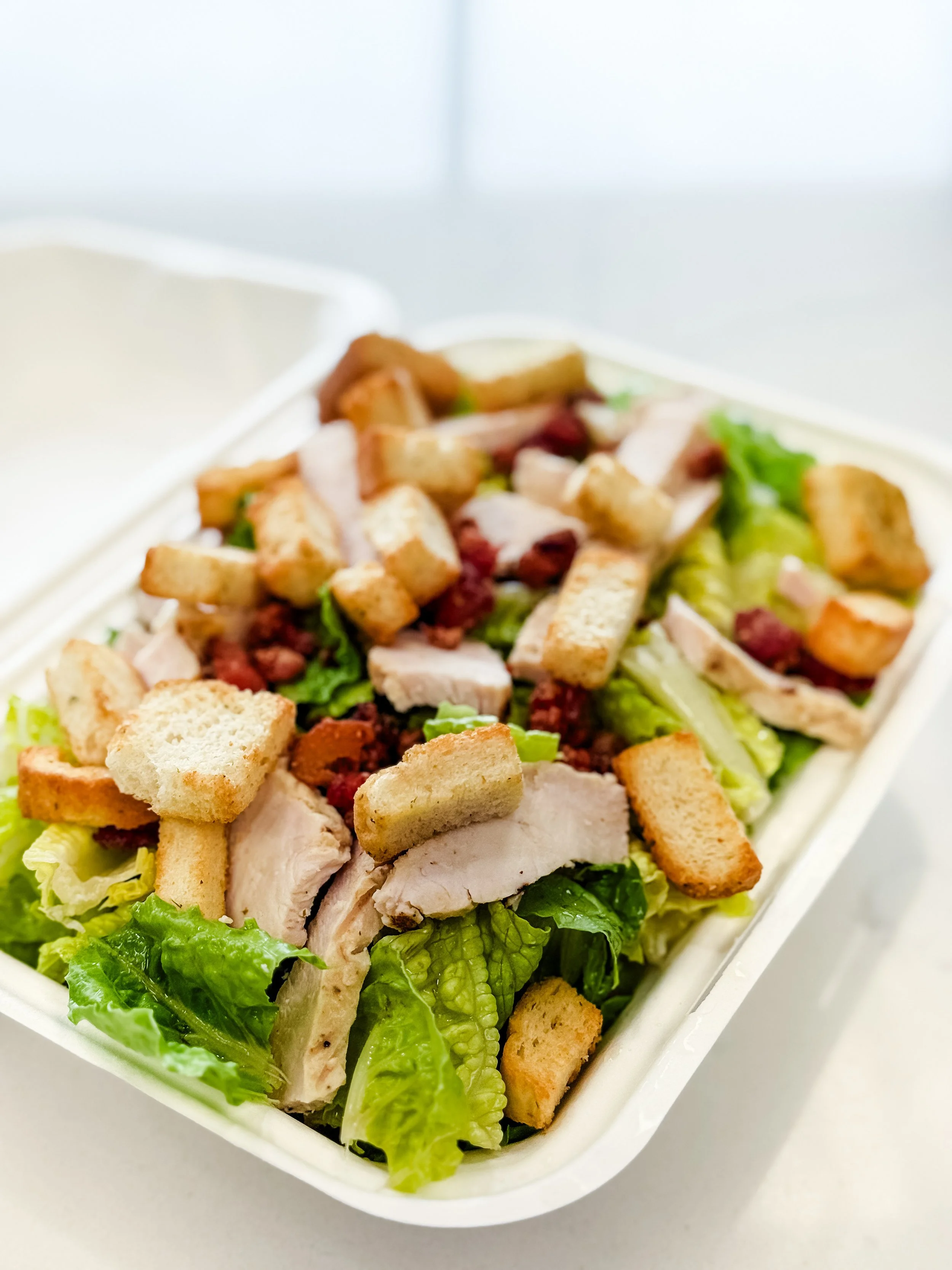 Caesar salad with chopped romaine lettuce, grilled chicken slices, croutons, and bacon bits in a takeout container.