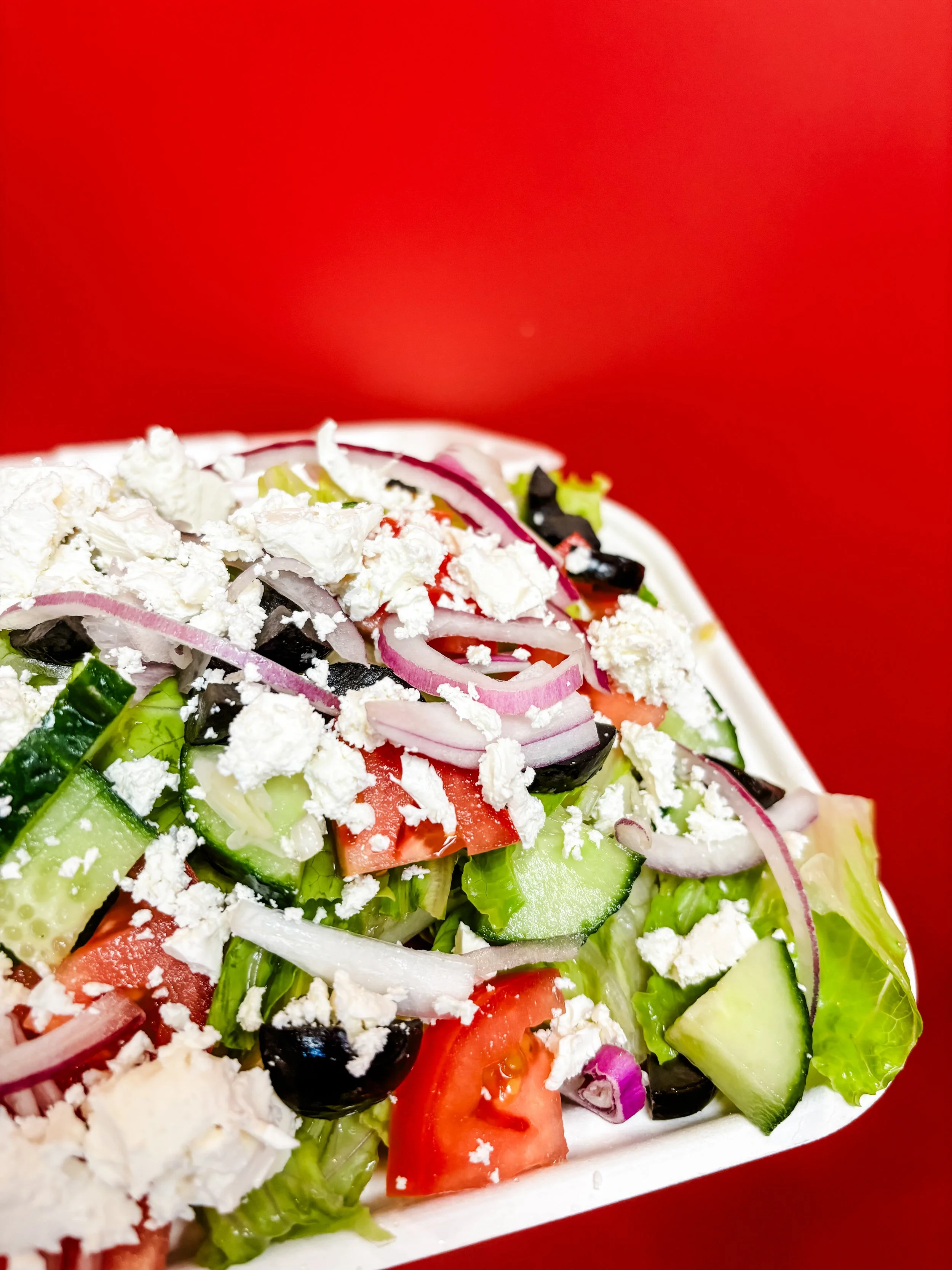Fresh vegetable salad with tomatoes, cucumbers, red onions, black olives, topped with crumbled feta cheese, placed on a white dish against a red background.