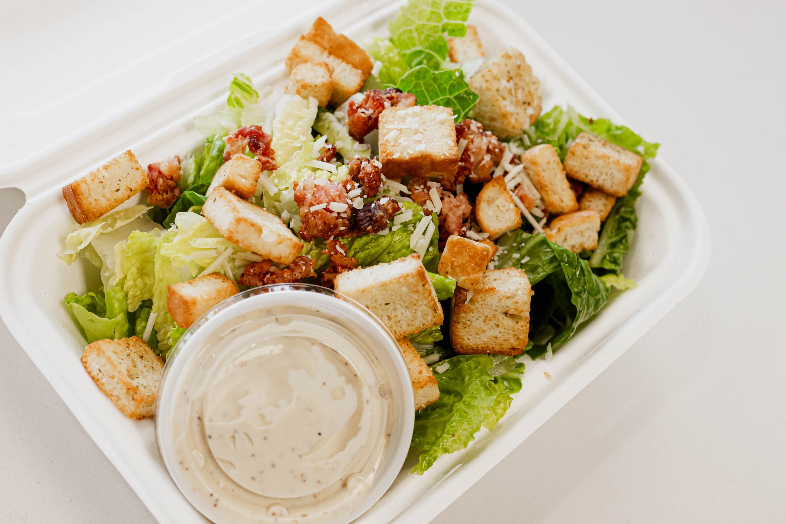 Caesar salad with croutons, bacon bits, shredded cheese, and a side of creamy dressing in a plastic container.