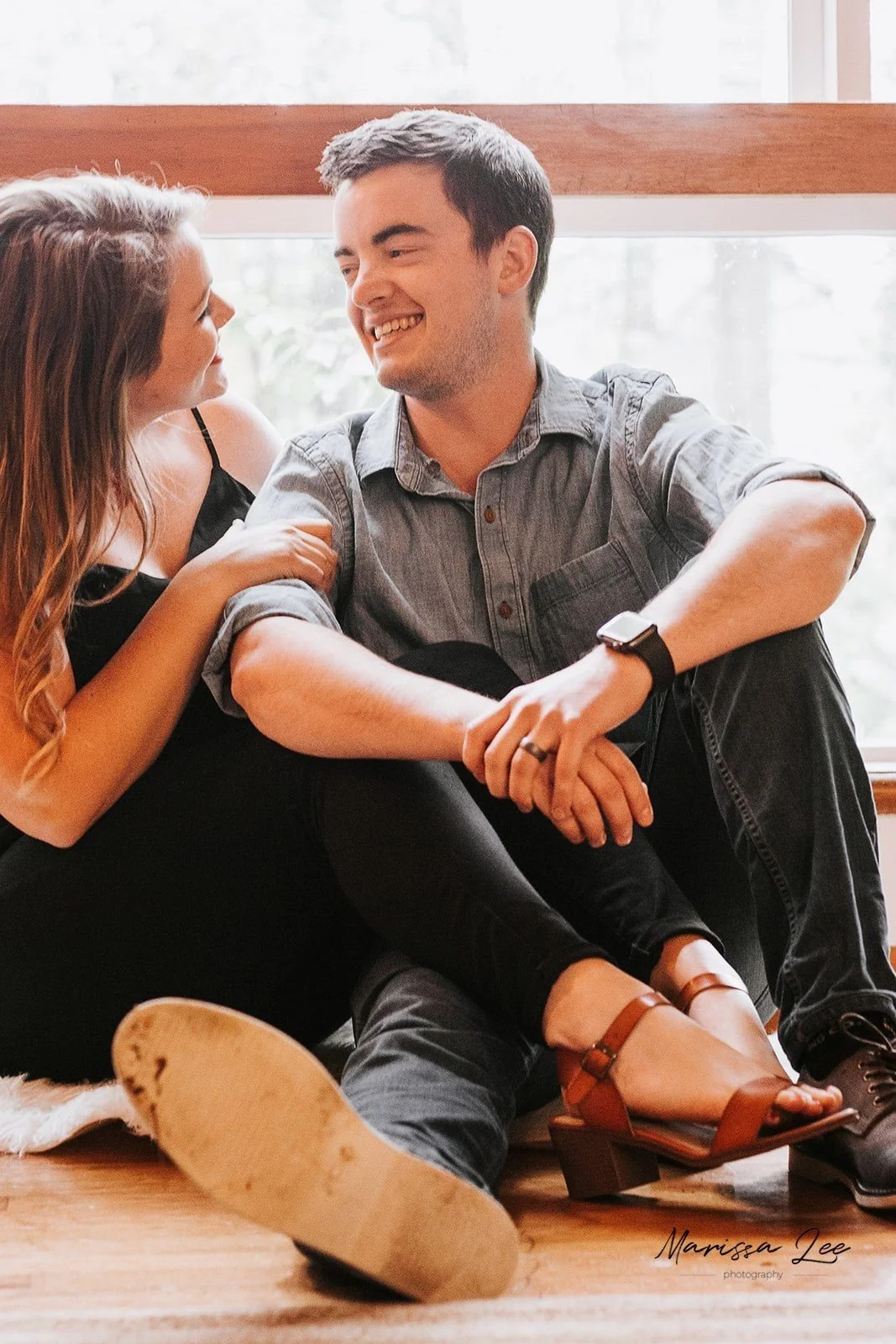A young couple sitting on a wooden floor near a large window, smiling and looking at each other. The woman has long wavy hair and is wearing a black top and black pants, while the man has short dark hair, is wearing a gray button-up shirt, dark pants