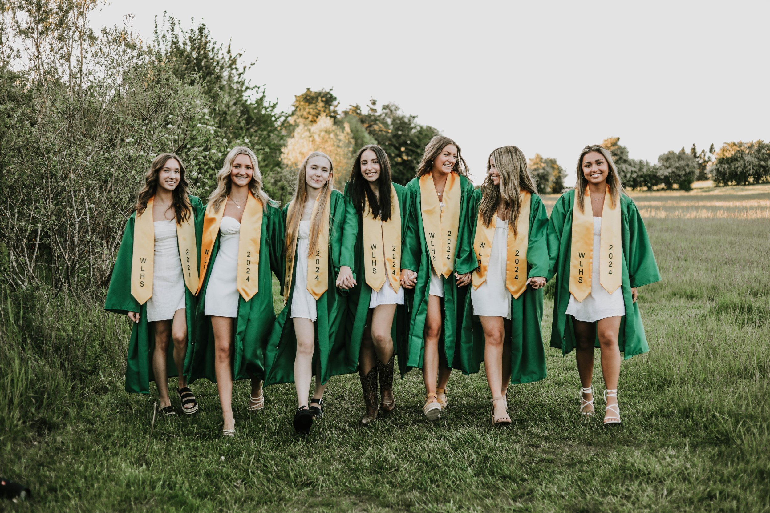 Group of seven young women in graduation gowns and sashes walking on grass with trees in the background.