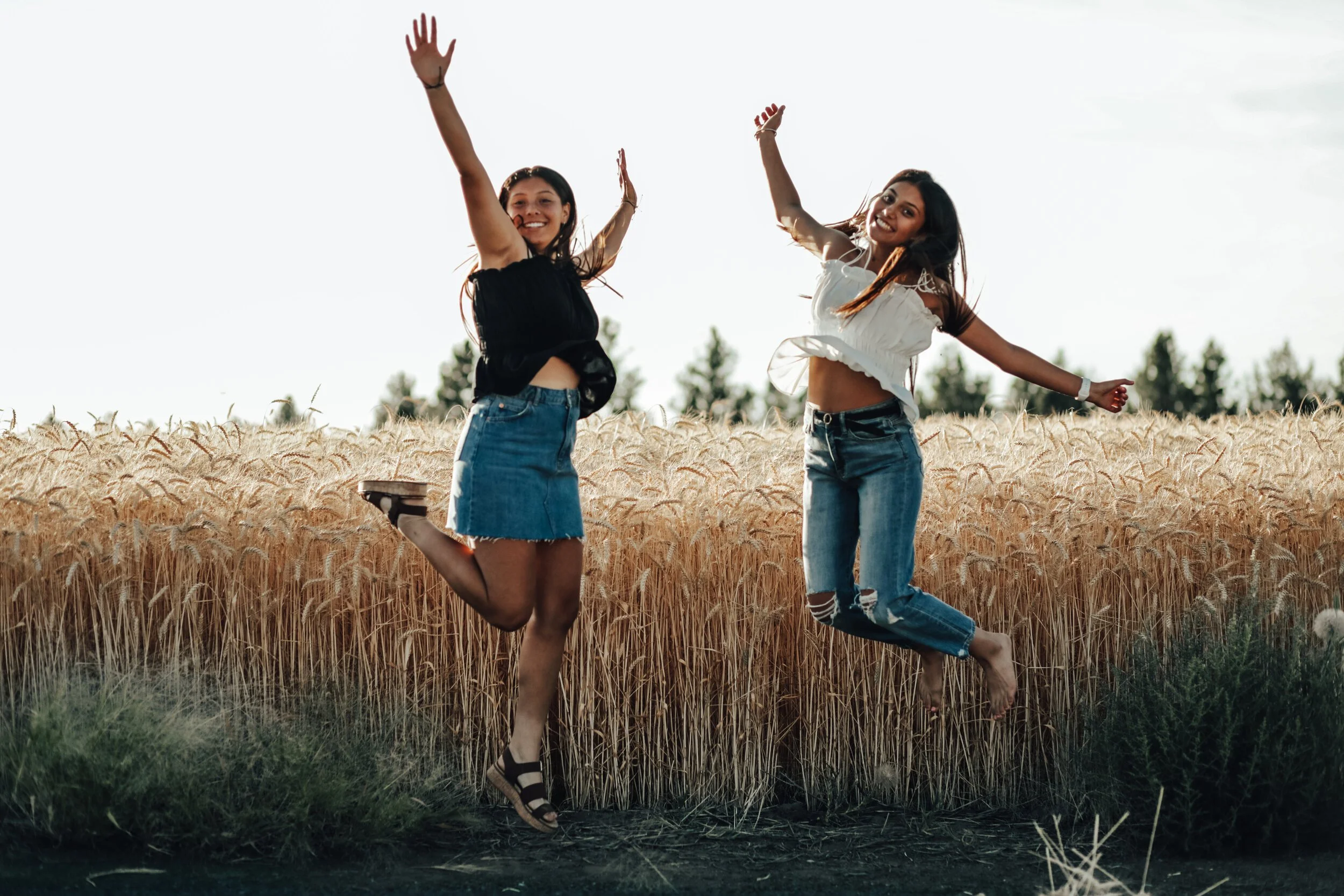 Two young women jumping and smiling in a wheat field during daylight with trees in the background.