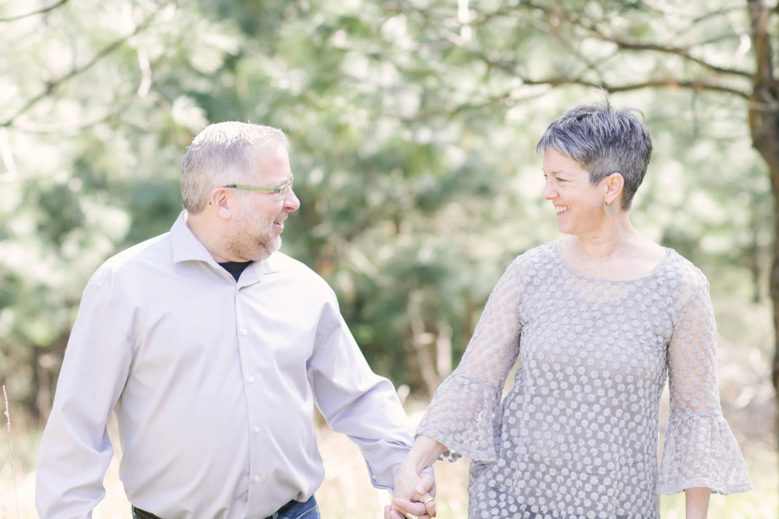 A smiling couple holding hands outdoors in a green, wooded setting.